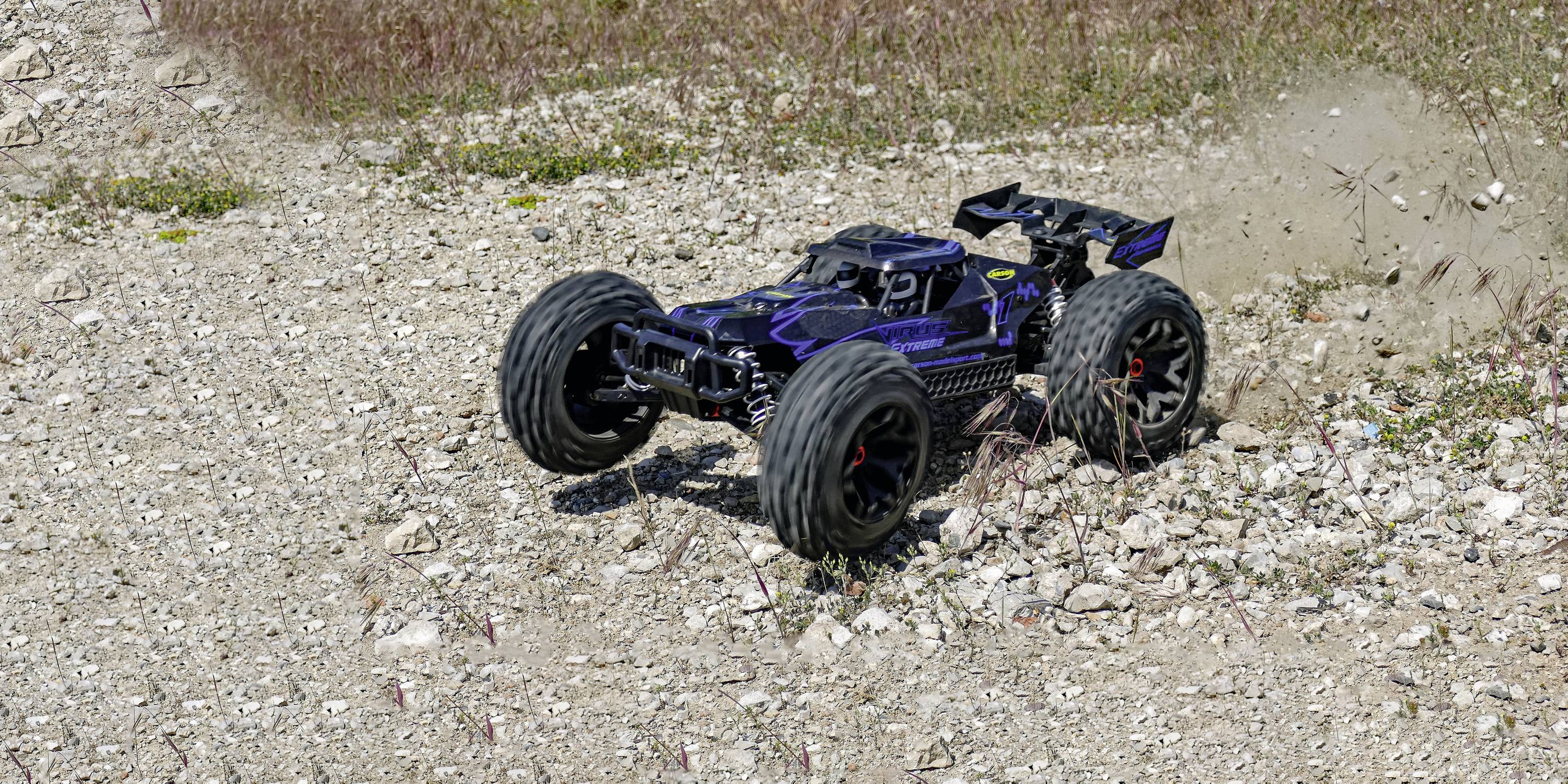 Une voiture télécommandée roule sur un terrain rocailleux, de la poussière soulevée indique son mouvement. De la végétation herbeuse est visible en arrière-plan.