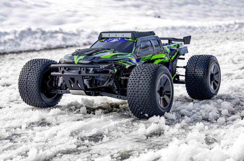Une voiture télécommandée avec un motif vert et noir roule dans la neige. Elle possède de grandes roues adaptées aux surfaces inégales et enneigées.