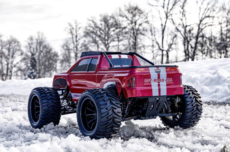 Un camion tout-terrain rouge avec de grandes roues est stationné dans un terrain enneigé. Des arbres dénudés et un ciel nuageux sont visibles en arrière-plan.