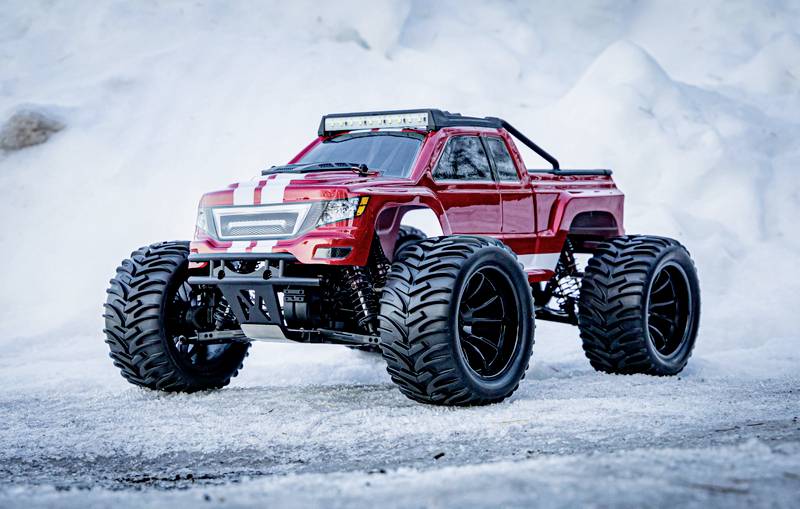 Un modèle de monster truck télécommandé rouge est posé sur un sol enneigé. De grandes roues, de la neige en arrière-plan.