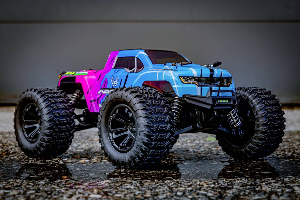 Une voiture télécommandée est posée sur un asphalte humide. Elle est bleue et rose avec de grandes roues tout-terrain à gros crampons, adaptées aux terrains accidentés.