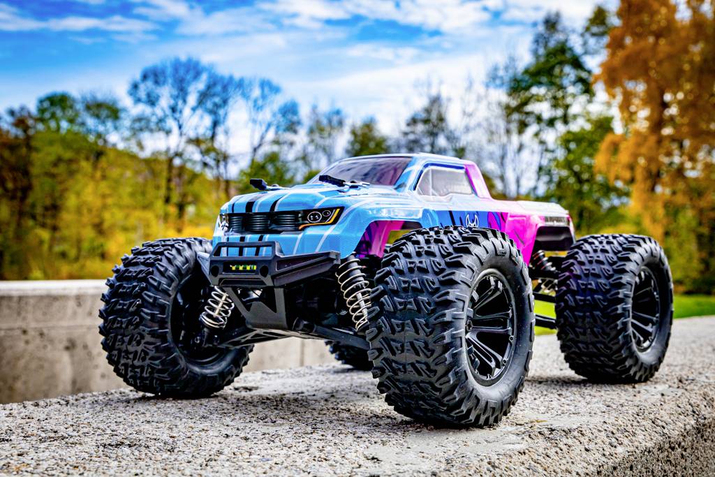 Une voiture télécommandée avec de grandes roues est garée sur une surface en béton. Des arbres aux feuilles d'automne sont visibles en arrière-plan.