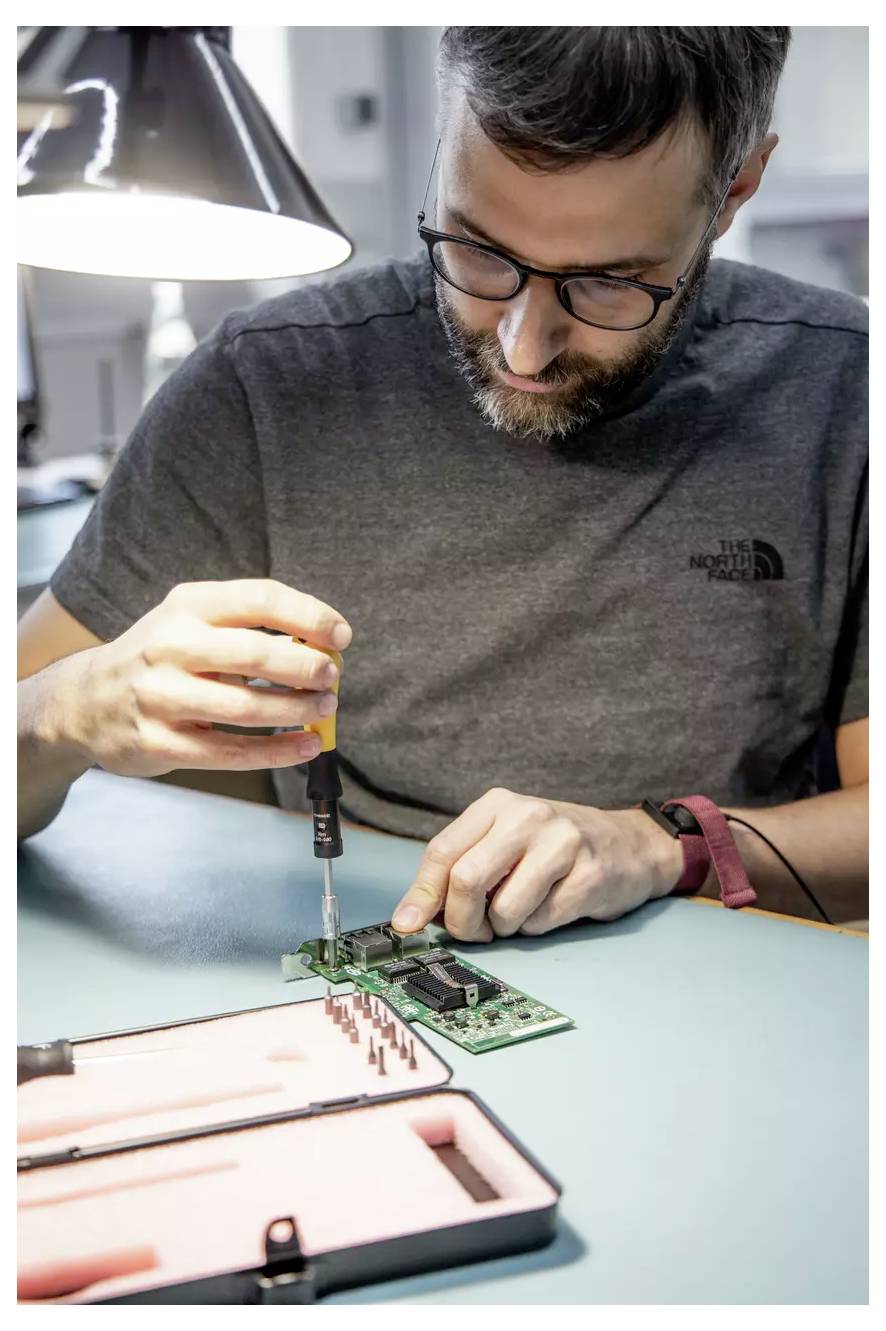Une personne portant des lunettes est concentrée sur l'utilisation d'un tournevis pour travailler sur une carte de circuit à un bureau, sous une lampe allumée. Une mallette à outils est ouverte à proximité.