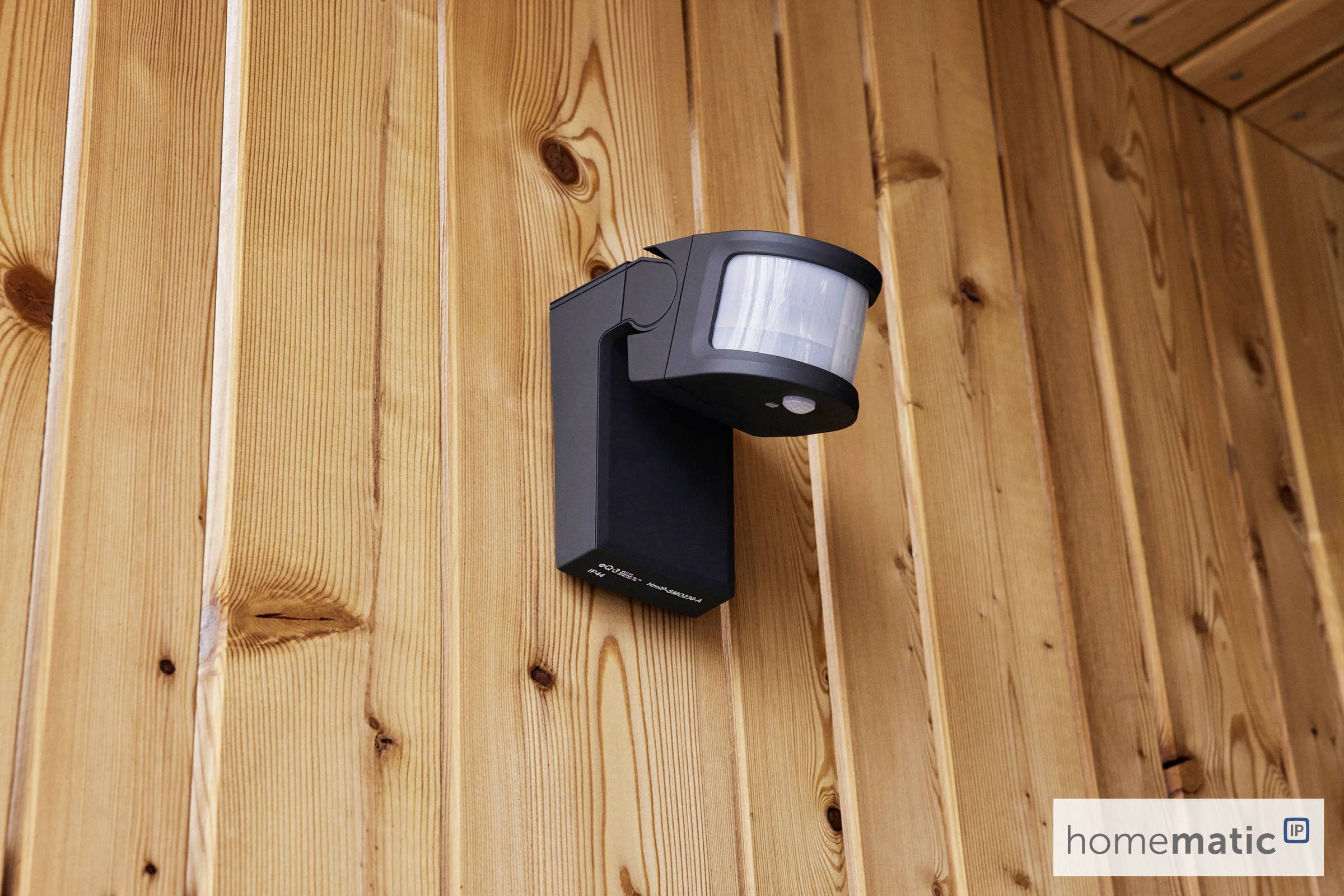 Capteur de mouvement noir monté sur un mur en bois. Le logo 'Homematic' est visible en bas à droite.