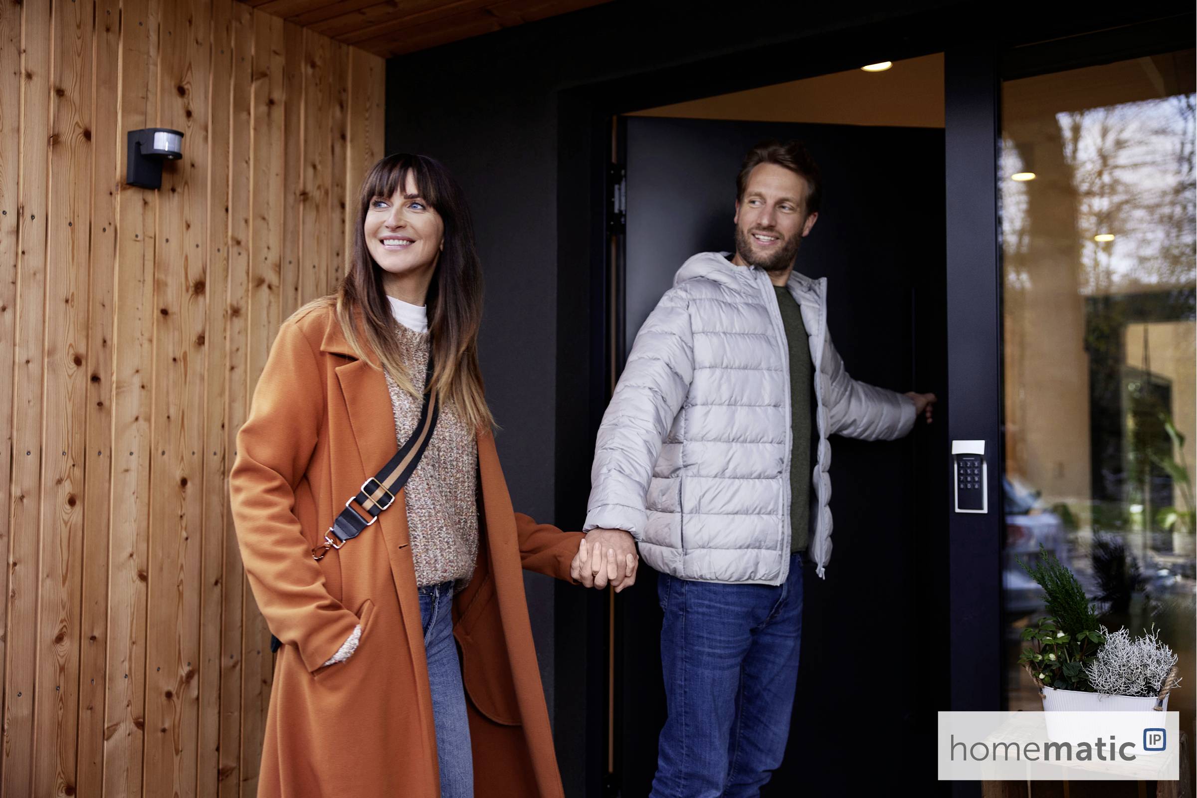 Un couple se tient par la main et quitte ensemble un bâtiment moderne. Tous deux sourient et portent des vêtements d'hiver.