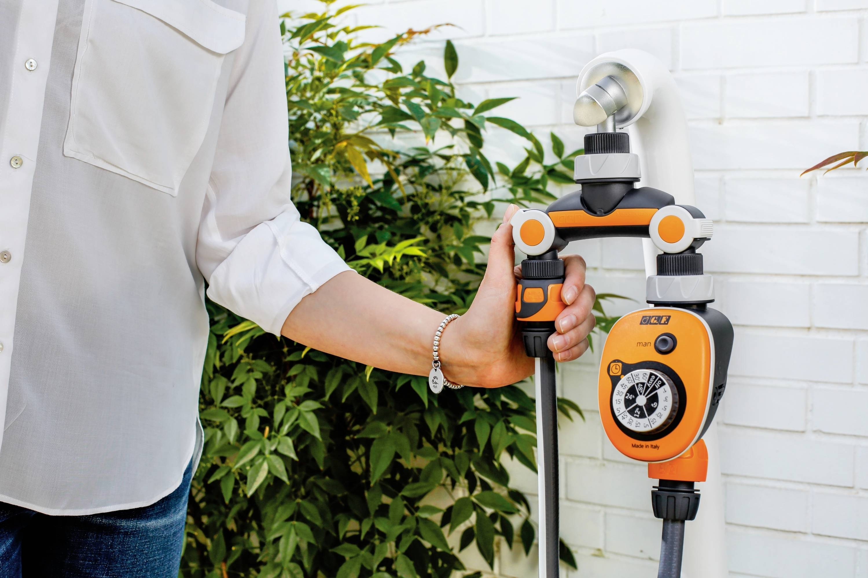 Une personne installe un programmateur d'arrosage orange et noir sur un robinet dans un jardin, entouré de plantes et d'un mur blanc.