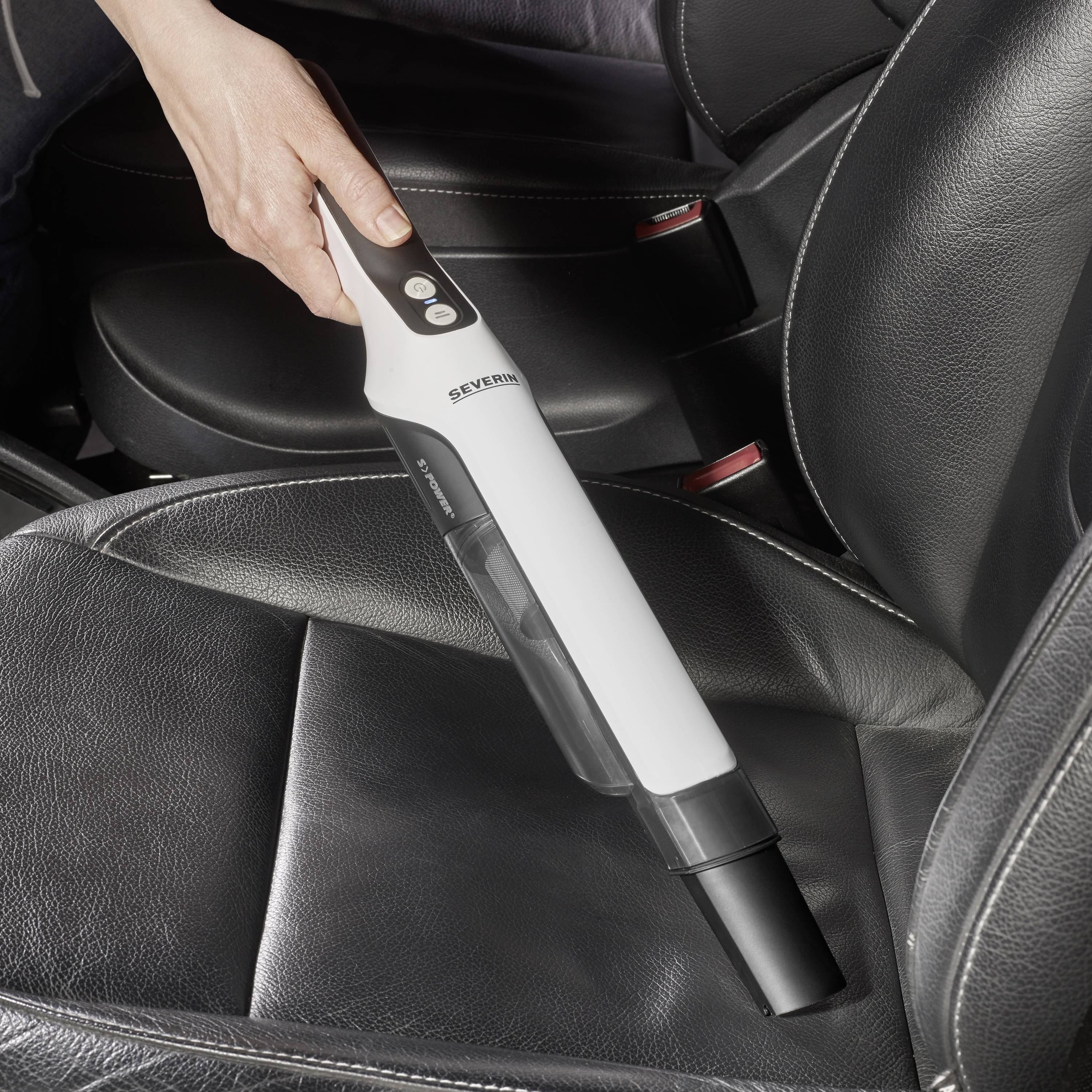 Une personne nettoie un siège de voiture avec un aspirateur à main blanc. Le siège est en cuir noir.