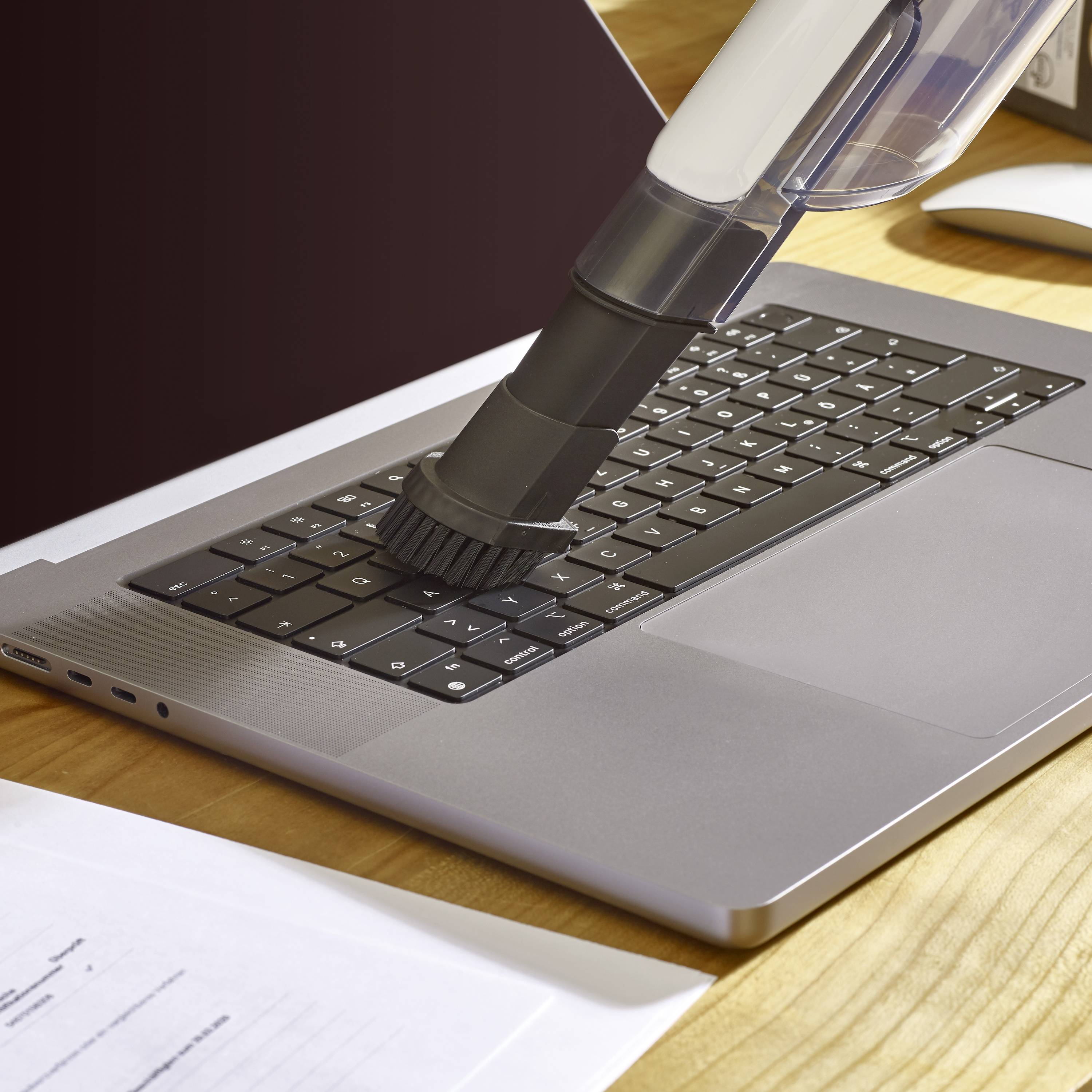 'Un embout d'aspirateur nettoie le clavier d'un ordinateur portable sur un bureau en bois, à côté de papiers et d'une souris d'ordinateur.'