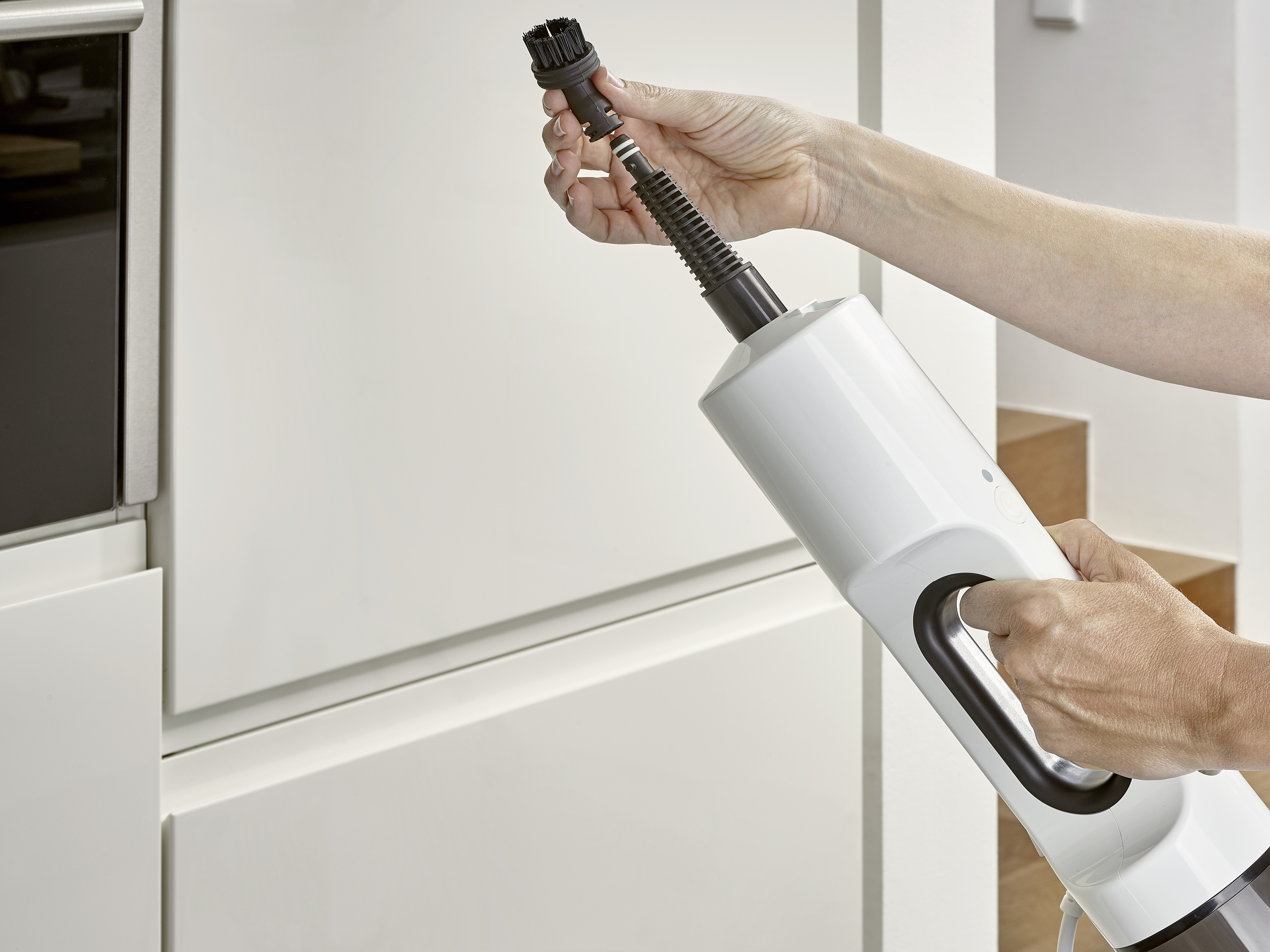 Une personne tient un aspirateur et fixe un embout brosse. Des armoires de cuisine sont visibles en arrière-plan.