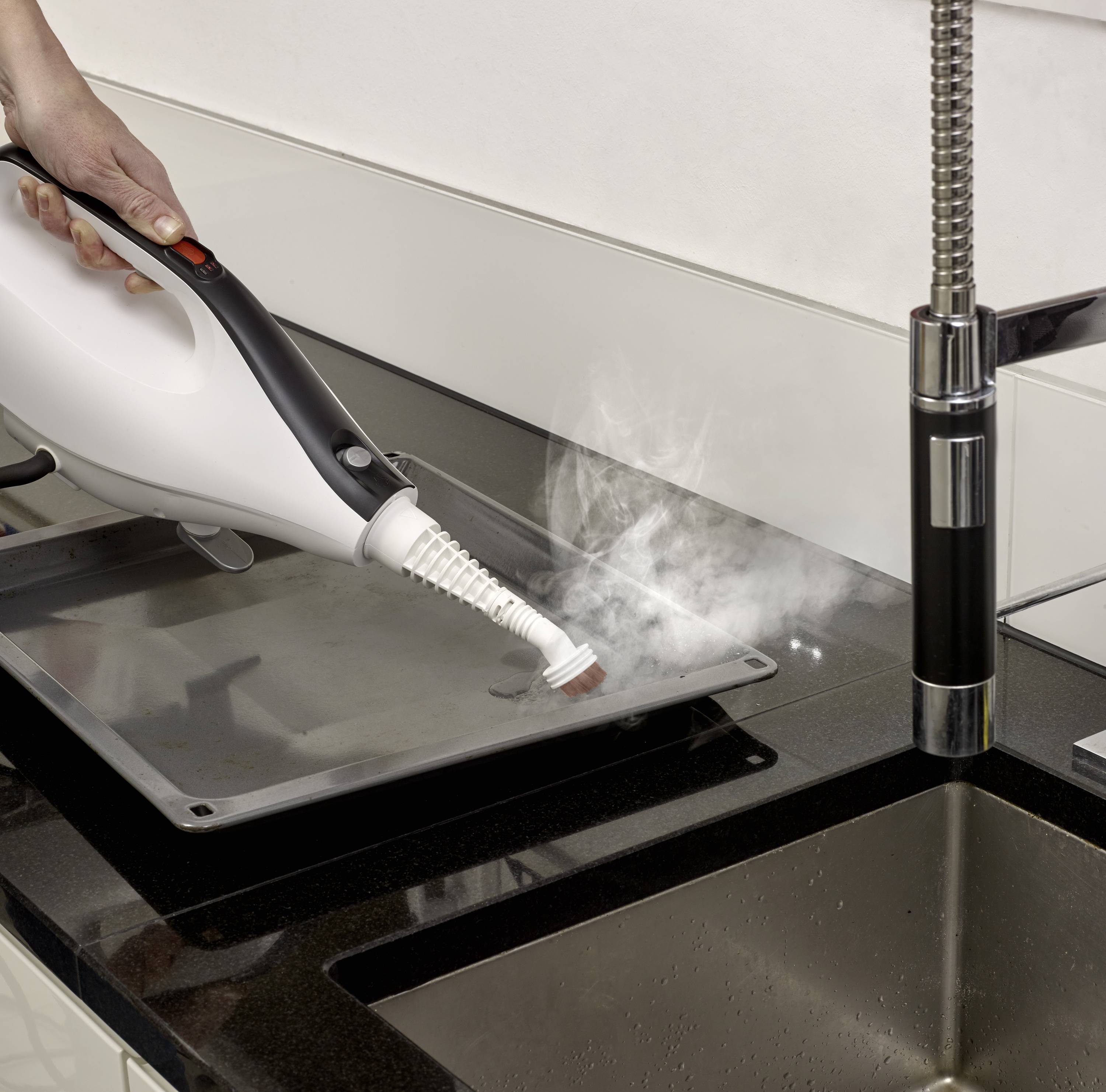 Une personne nettoie une plaque de cuisson avec un nettoyeur vapeur. La vapeur élimine les salissures de la surface près d'un évier en acier inoxydable.