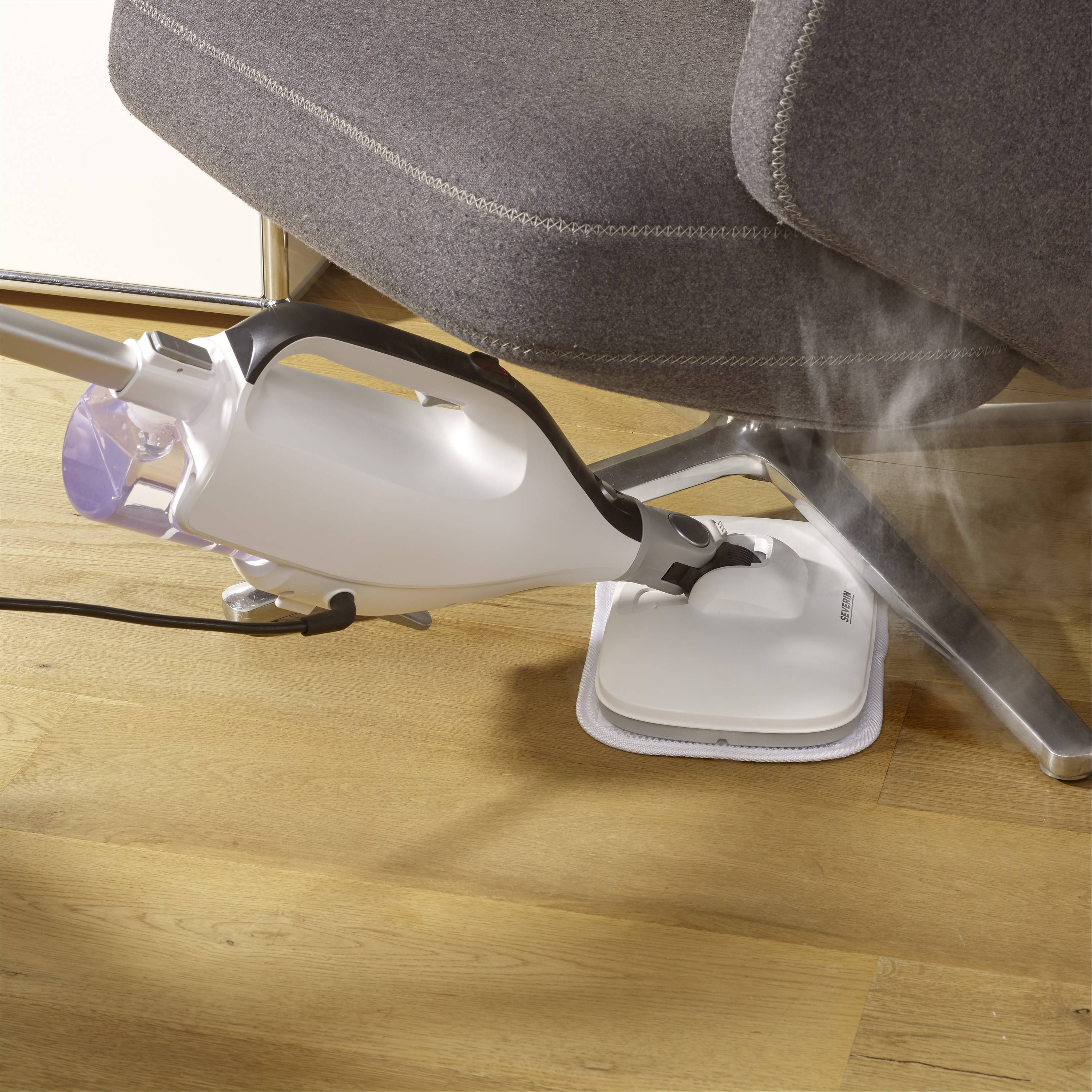 Nettoyeur vapeur générant de la vapeur sur un parquet en bois sous une chaise grise. Nettoie soigneusement le sol avec de la vapeur d'eau visible.
