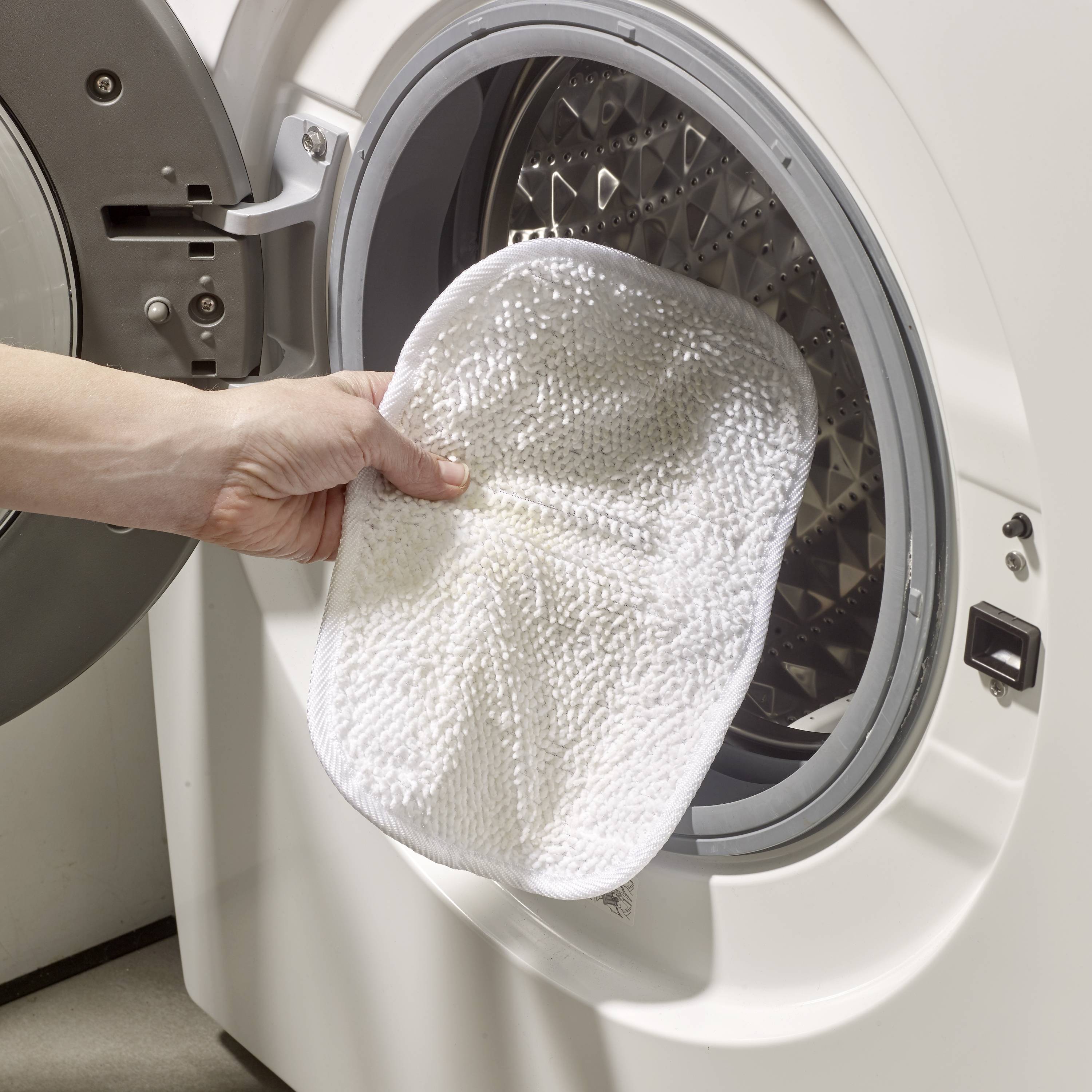 Une main dépose un chiffon sale dans une machine à laver ouverte. La scène illustre une tâche ménagère quotidienne.