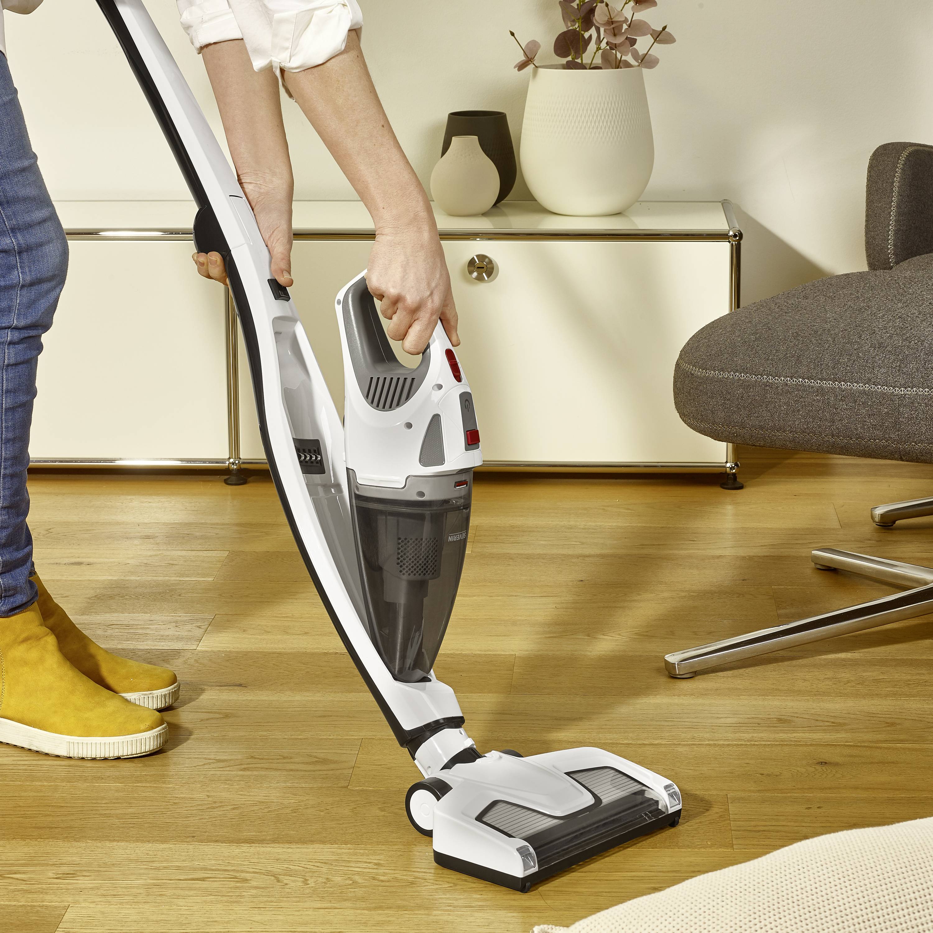 Une personne aspire un sol en bois avec un aspirateur sans fil dans une pièce chaleureuse ; une commode se trouve en arrière-plan.