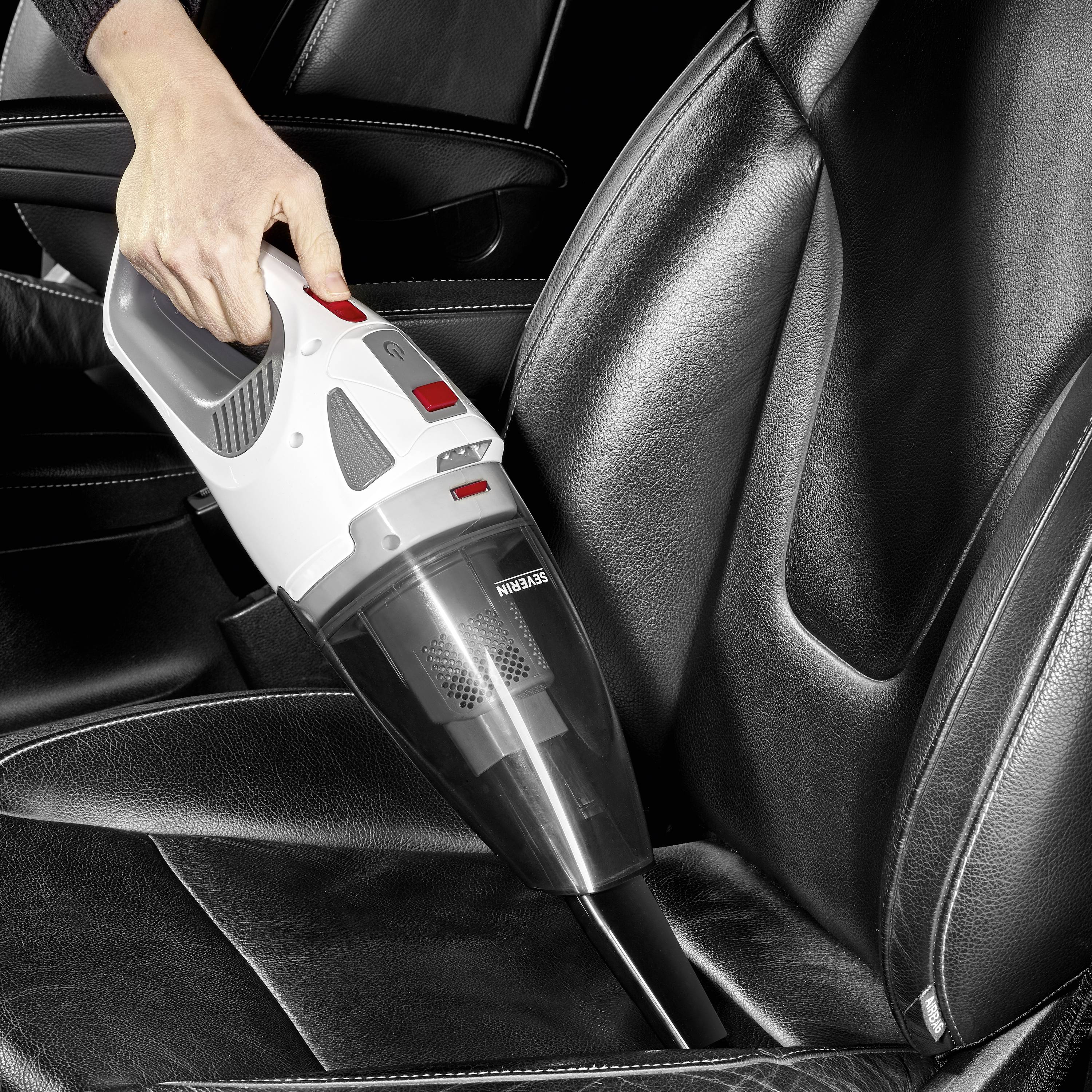 Une personne nettoie un siège de voiture avec un aspirateur à main. Le siège est en cuir noir et l'appareil est blanc et noir.