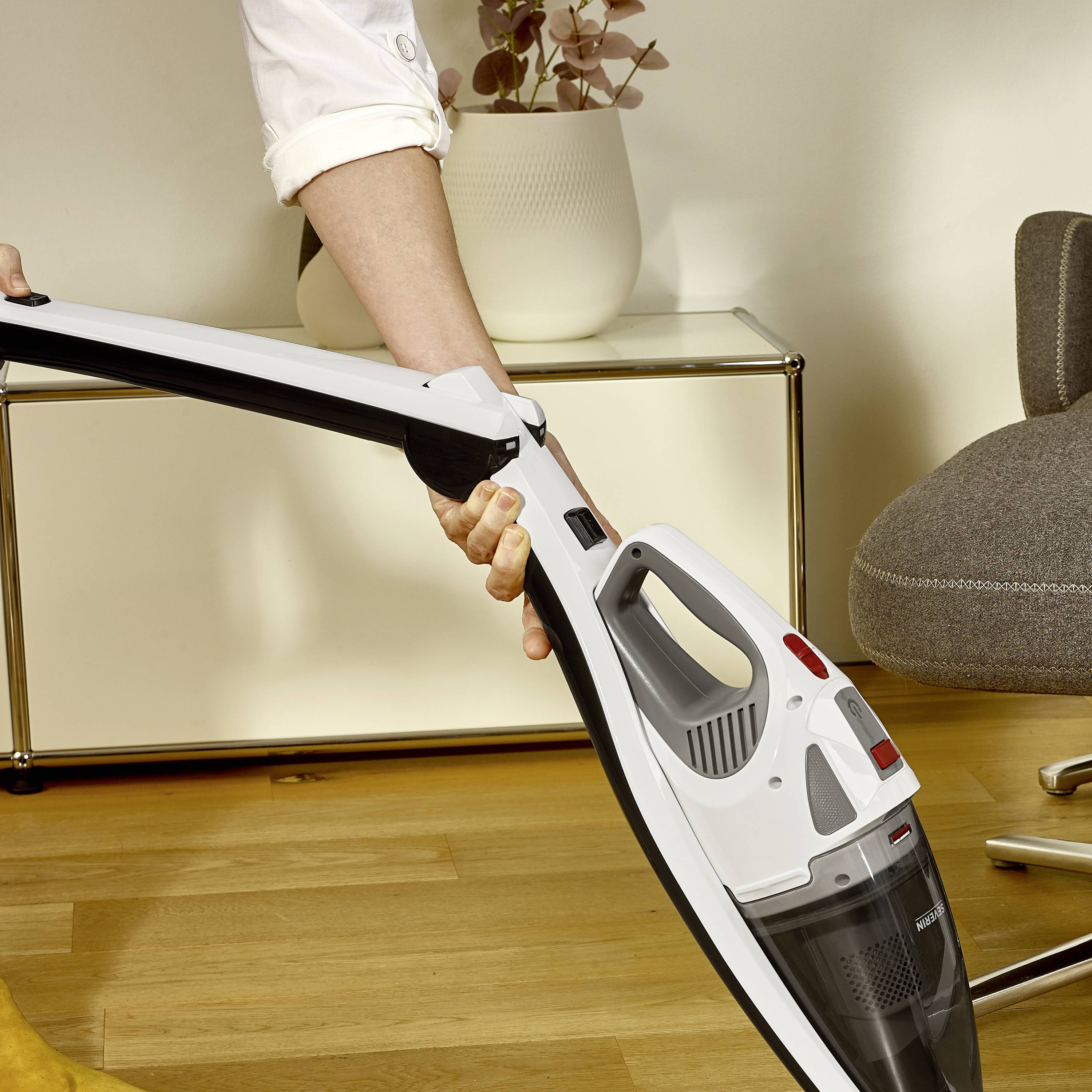 Une personne tient un aspirateur à main au-dessus d'un sol en bois. À l'arrière-plan, on aperçoit une petite table avec un vase et un fauteuil.