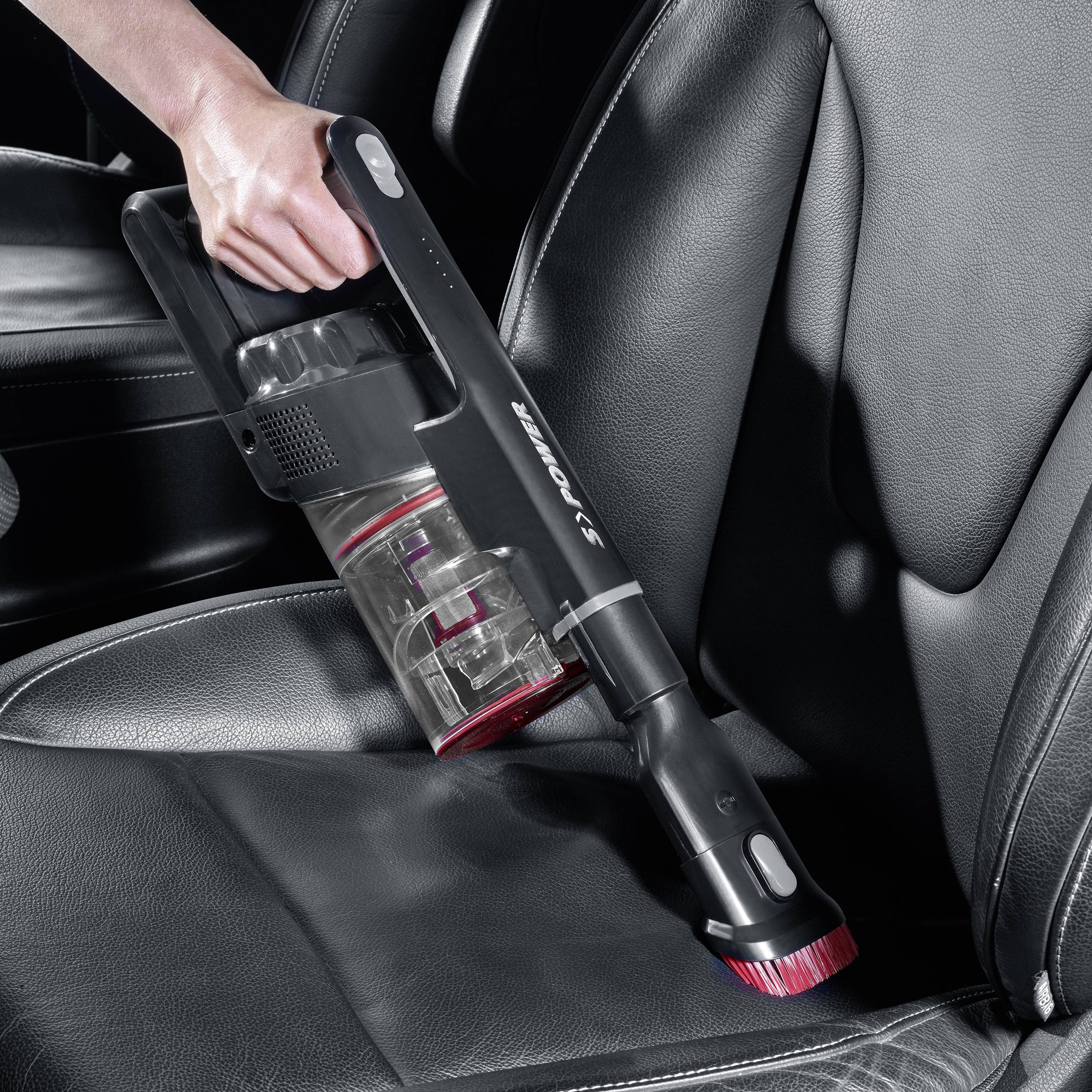 Une personne utilise un aspirateur portable pour nettoyer un siège de voiture. L'aspirateur a un réservoir à poussière transparent.