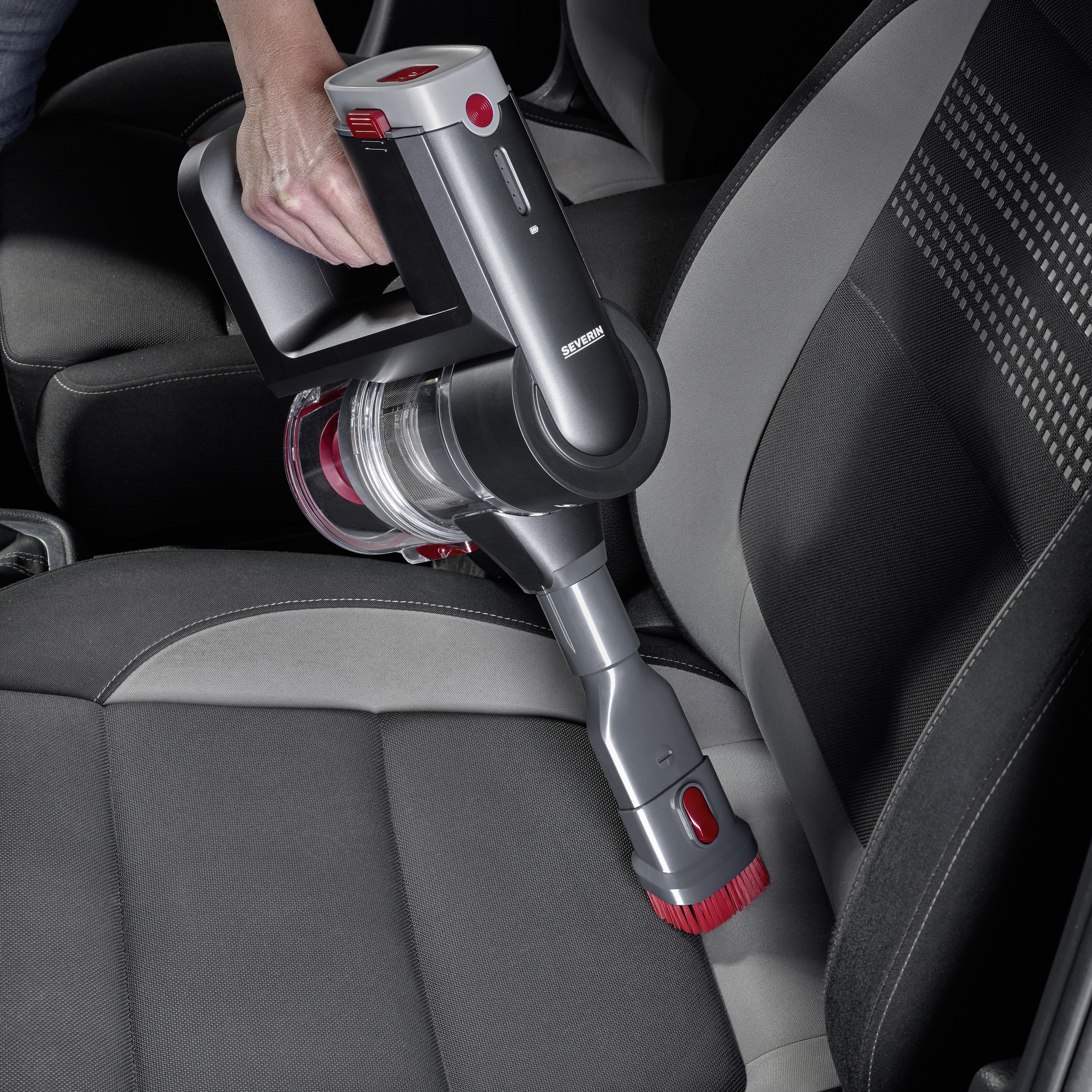 Une personne aspire le siège d'une voiture avec un aspirateur à main sans fil. L'aspirateur est compact et maniable.