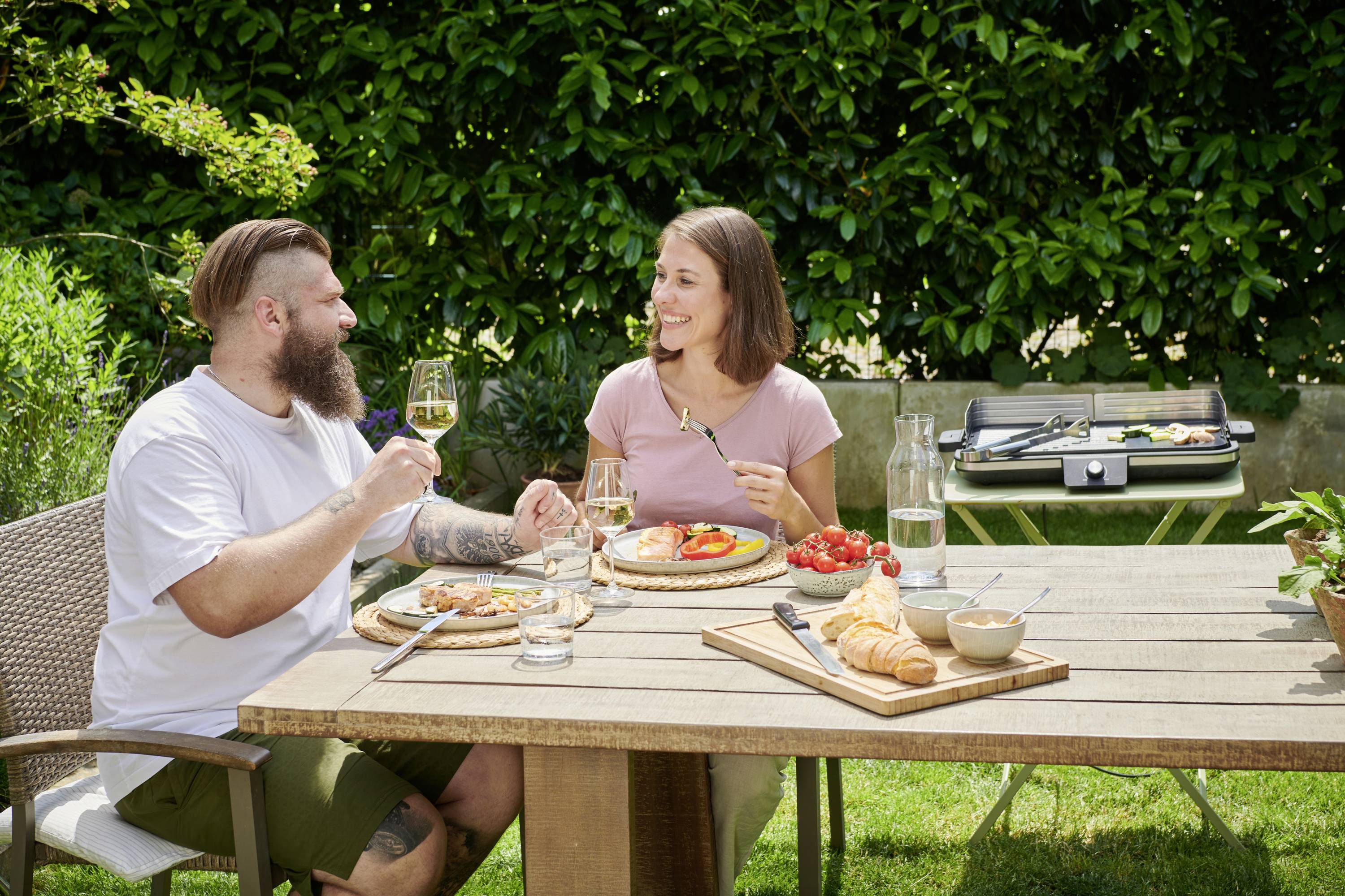 Un homme et une femme sont assis à une table dans un jardin et mangent. Ils sourient et lèvent leurs verres pour porter un toast. Un barbecue est visible en arrière-plan.