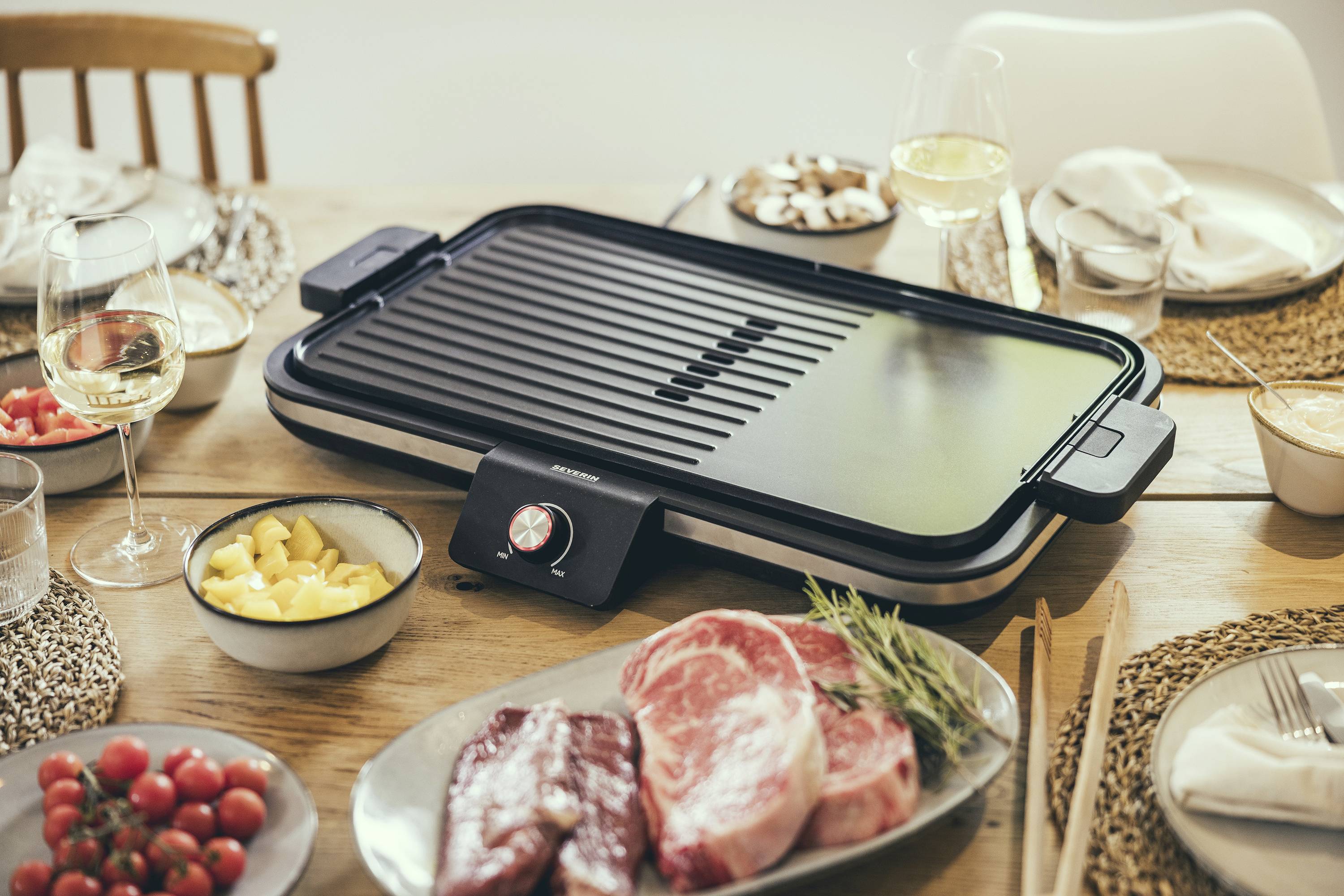 Grill électrique sur une table dressée avec de la viande crue, des fruits et des verres de vin blanc. Préparatifs pour un repas partagé.