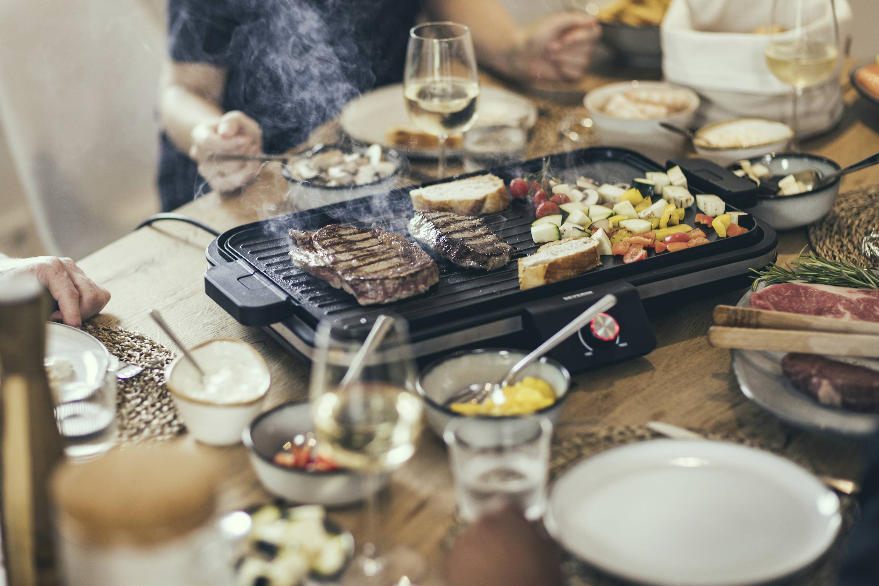 Un grill de table au centre d'une table dressée avec de la viande et des légumes. Des personnes sont assises autour de la table, il y a du pain et des boissons.