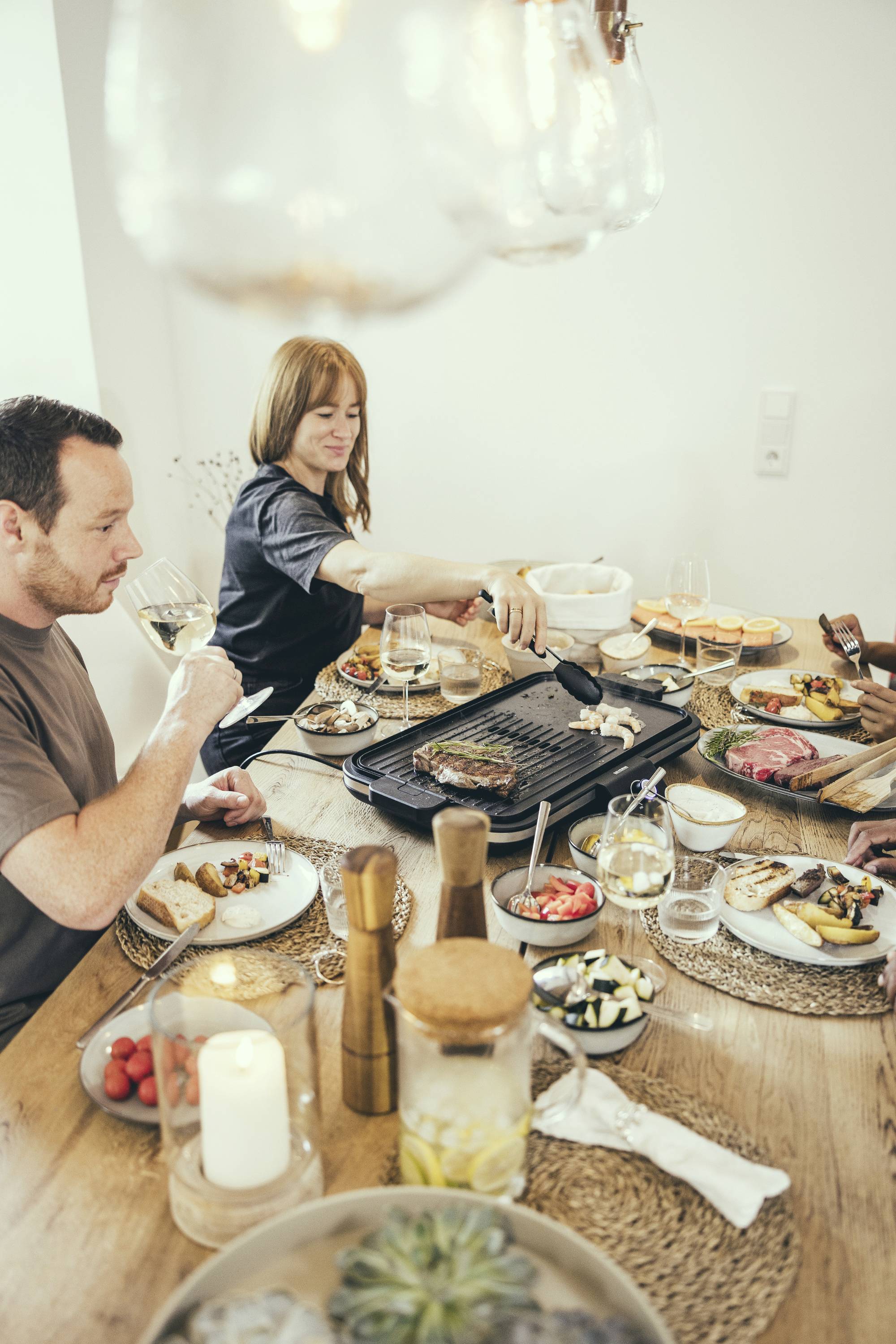 Un homme et une femme sont assis à une table dressée pendant qu'ils mangent. La femme fait griller des aliments sur une plaque de grill, tandis que l'homme tient un verre de vin.