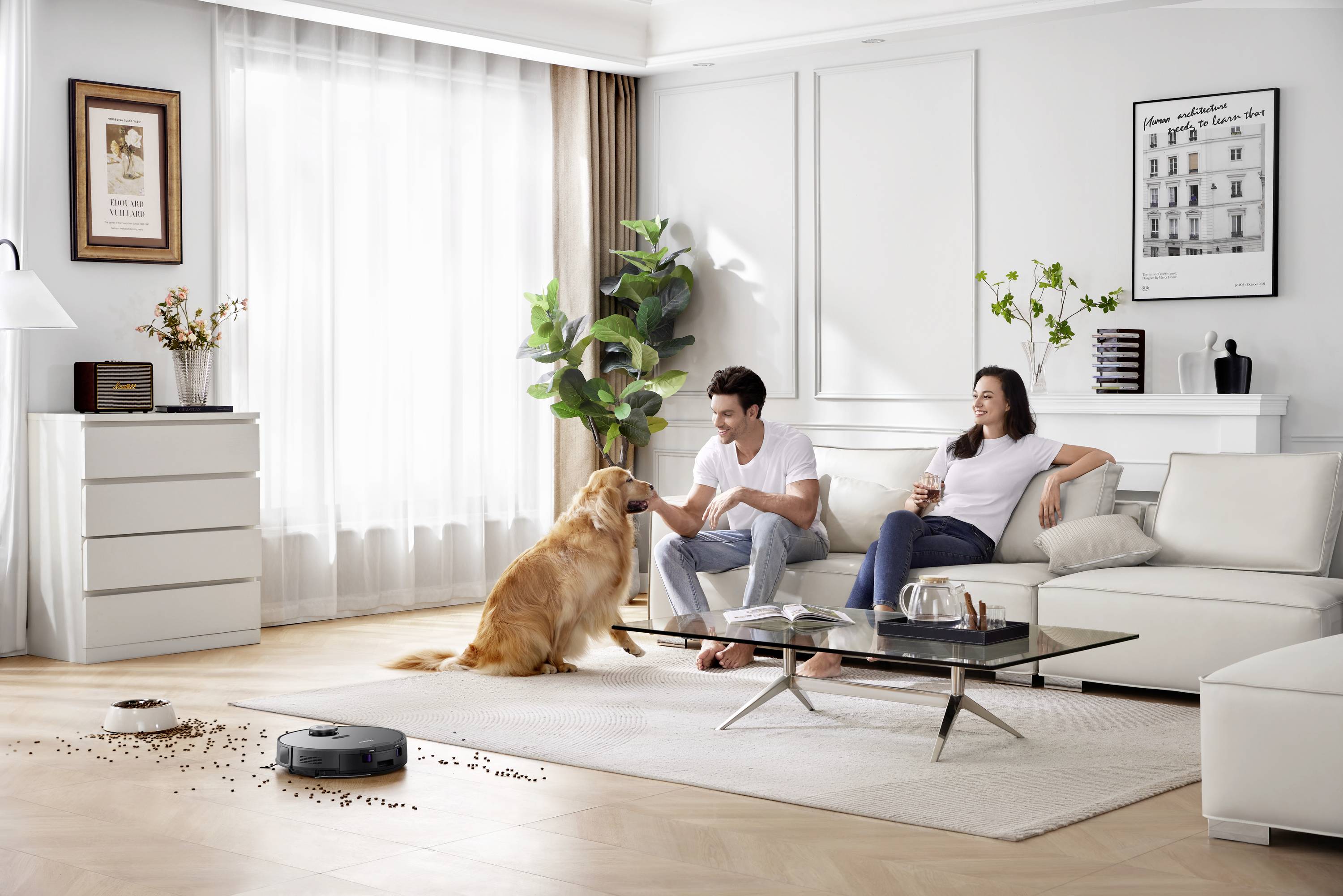 Un couple est assis sur un canapé dans un salon moderne, tandis qu'un chien est assis en face d'eux. Un aspirateur robot est en marche.