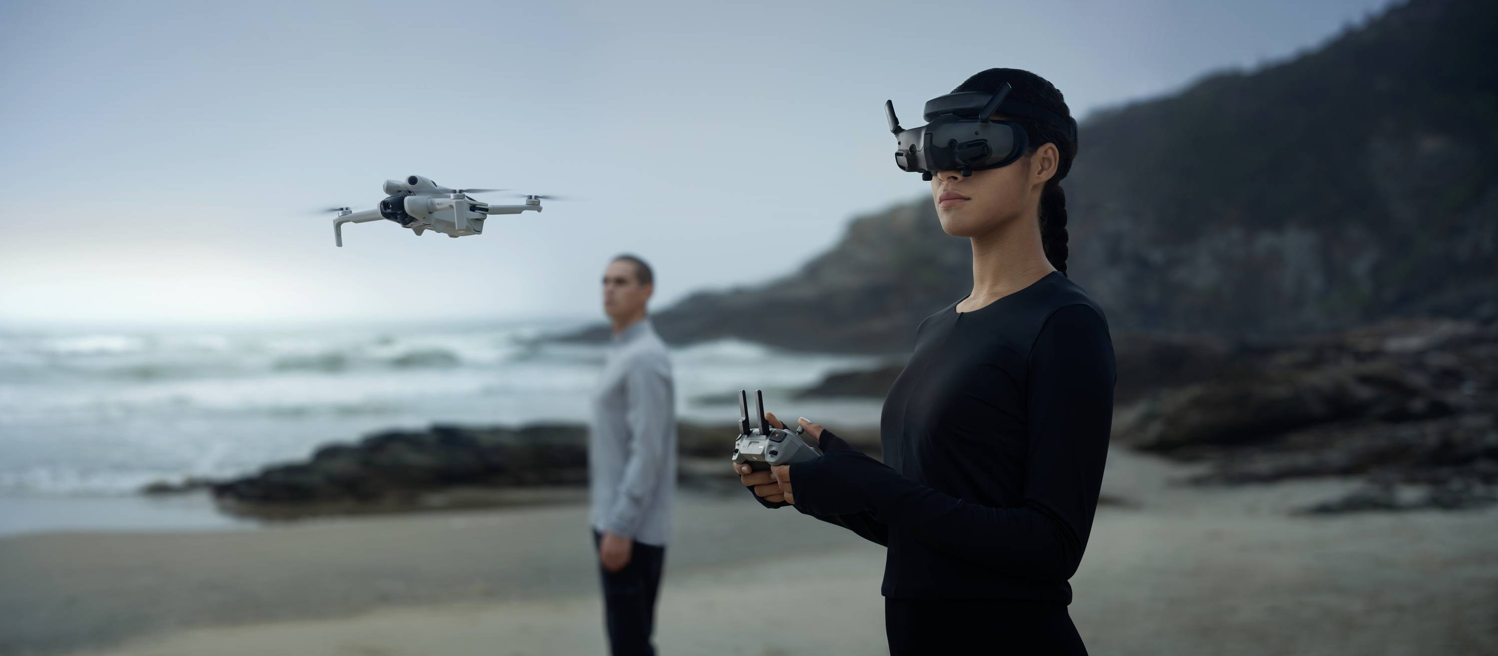 Une femme pilote un drone avec un casque de réalité virtuelle sur une plage, tandis qu'un homme se tient en arrière-plan.