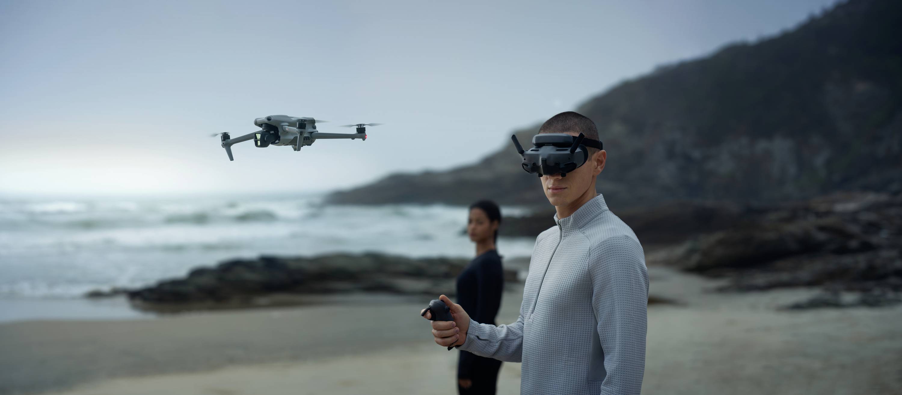 Deux personnes portant des lunettes de réalité virtuelle pilotent un drone sur une plage au coucher du soleil. Le drone plane au-dessus du sable, avec la mer en arrière-plan.