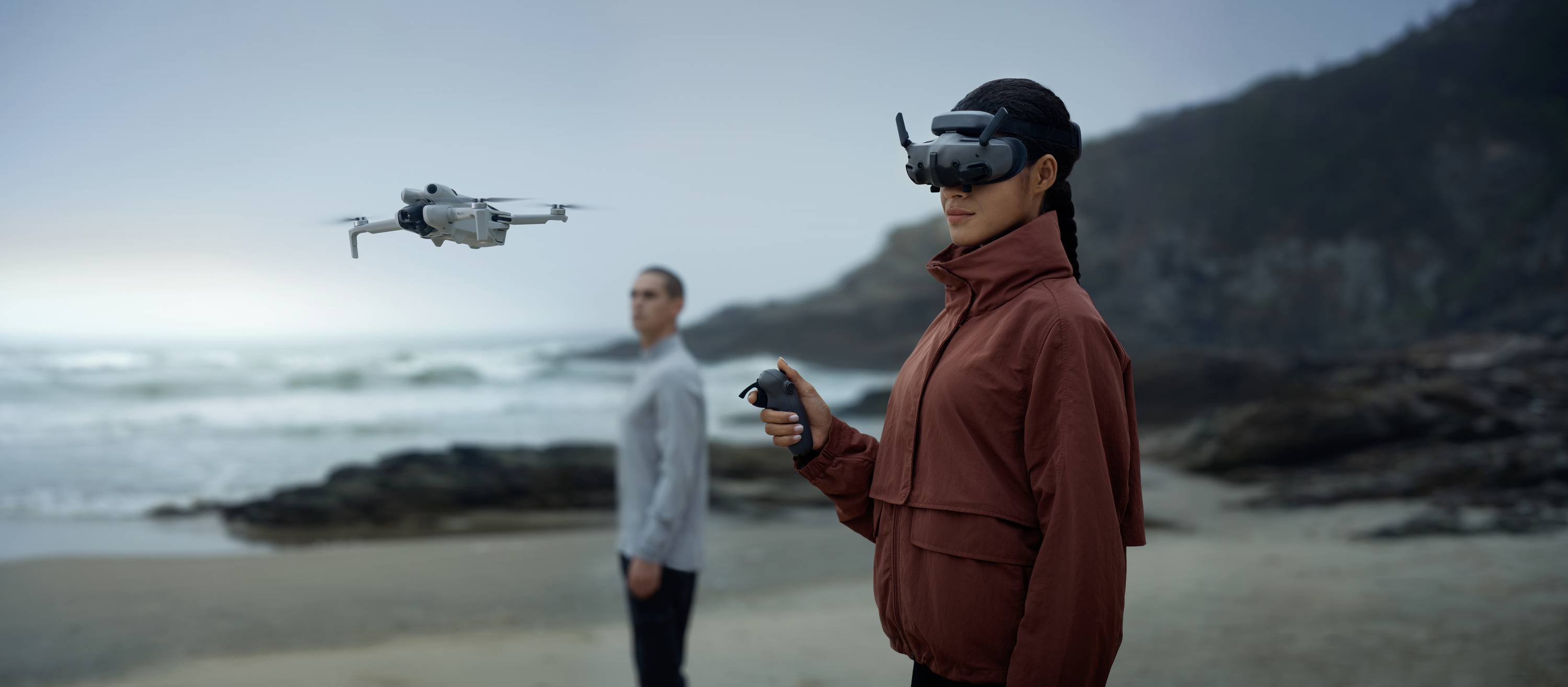 Une personne portant un casque de réalité virtuelle pilote un drone sur la plage. Une autre personne se tient en arrière-plan et regarde vers la mer.