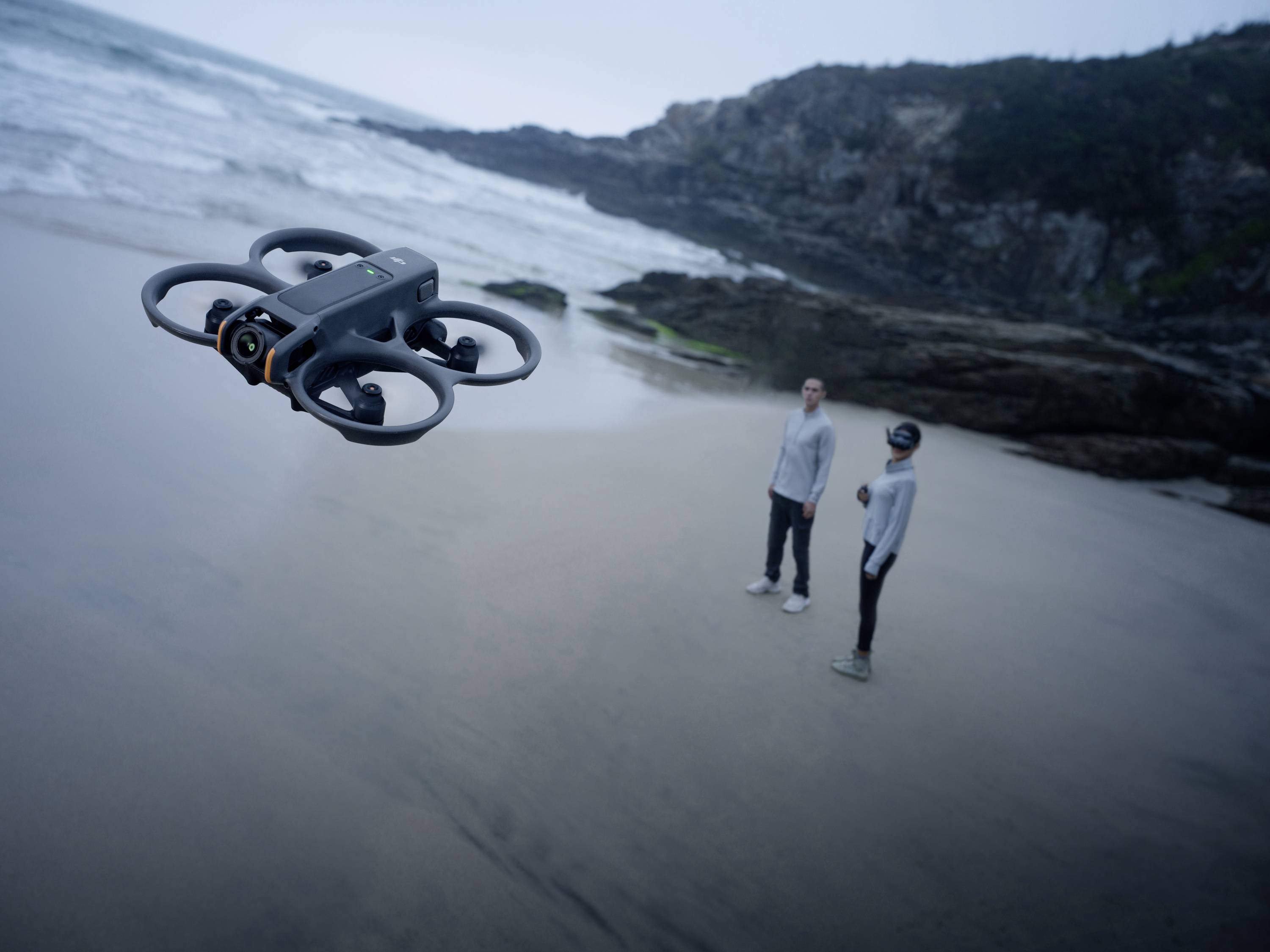 Un drone survole une plage, tandis que deux personnes se tiennent sur le rivage et regardent en direction du drone. Des rochers sont visibles en arrière-plan.