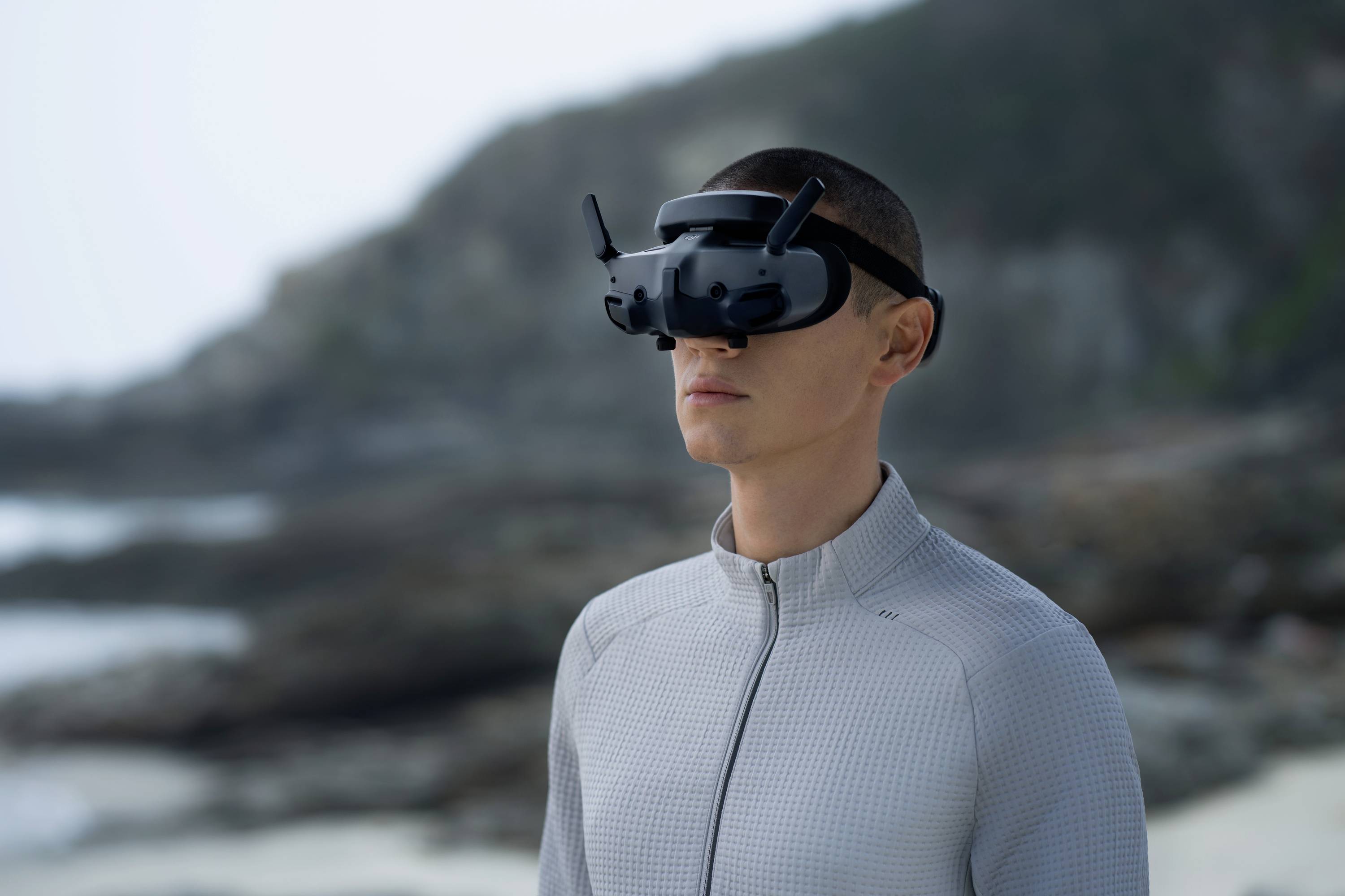 Une personne portant un casque de réalité virtuelle se tient sur la plage, le regard dirigé vers l'avant. Des rochers et la mer sont visibles en arrière-plan.