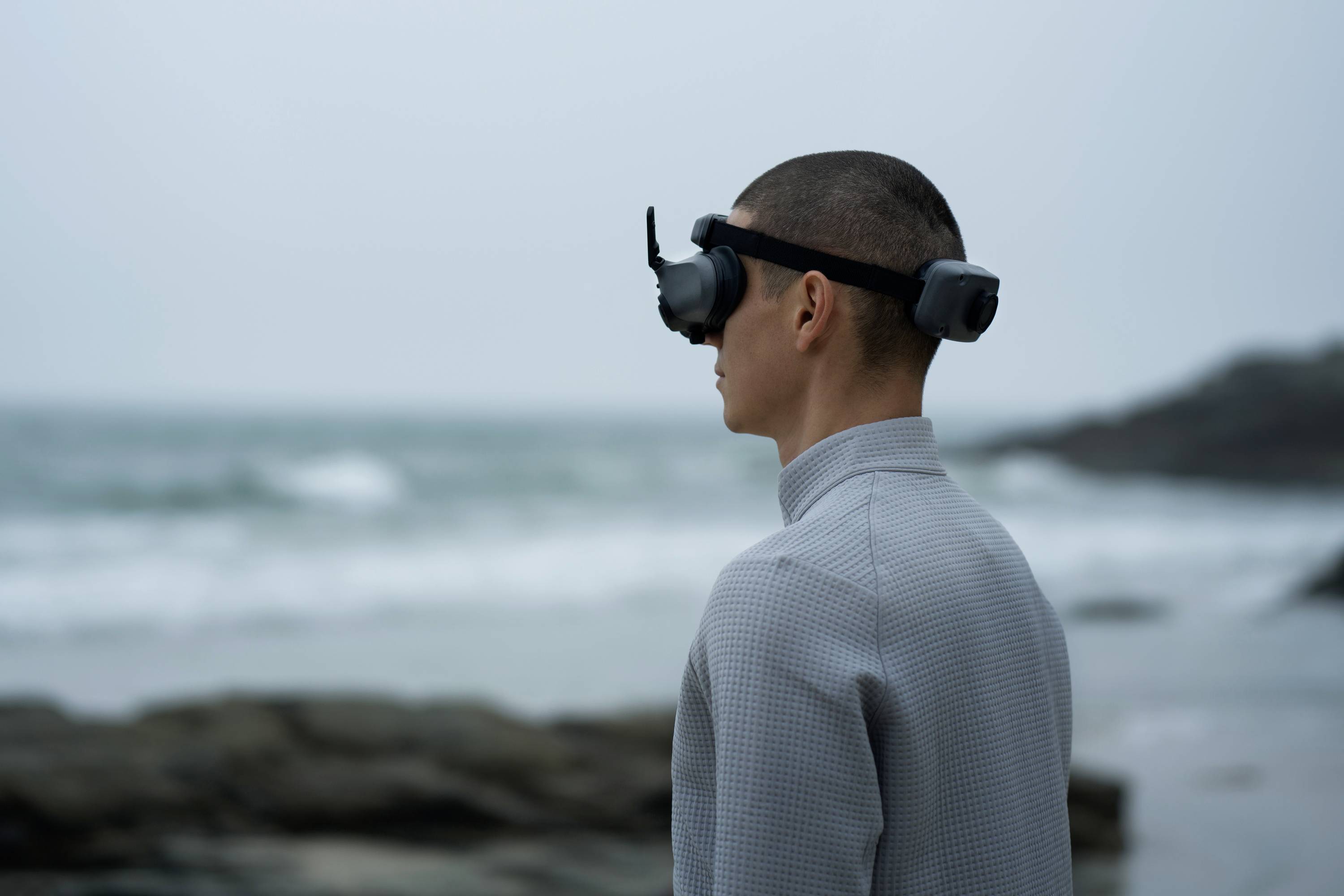 Une personne se tient au bord de la mer, portant un casque de réalité virtuelle. L'arrière-plan montre des vagues et un ciel nuageux.