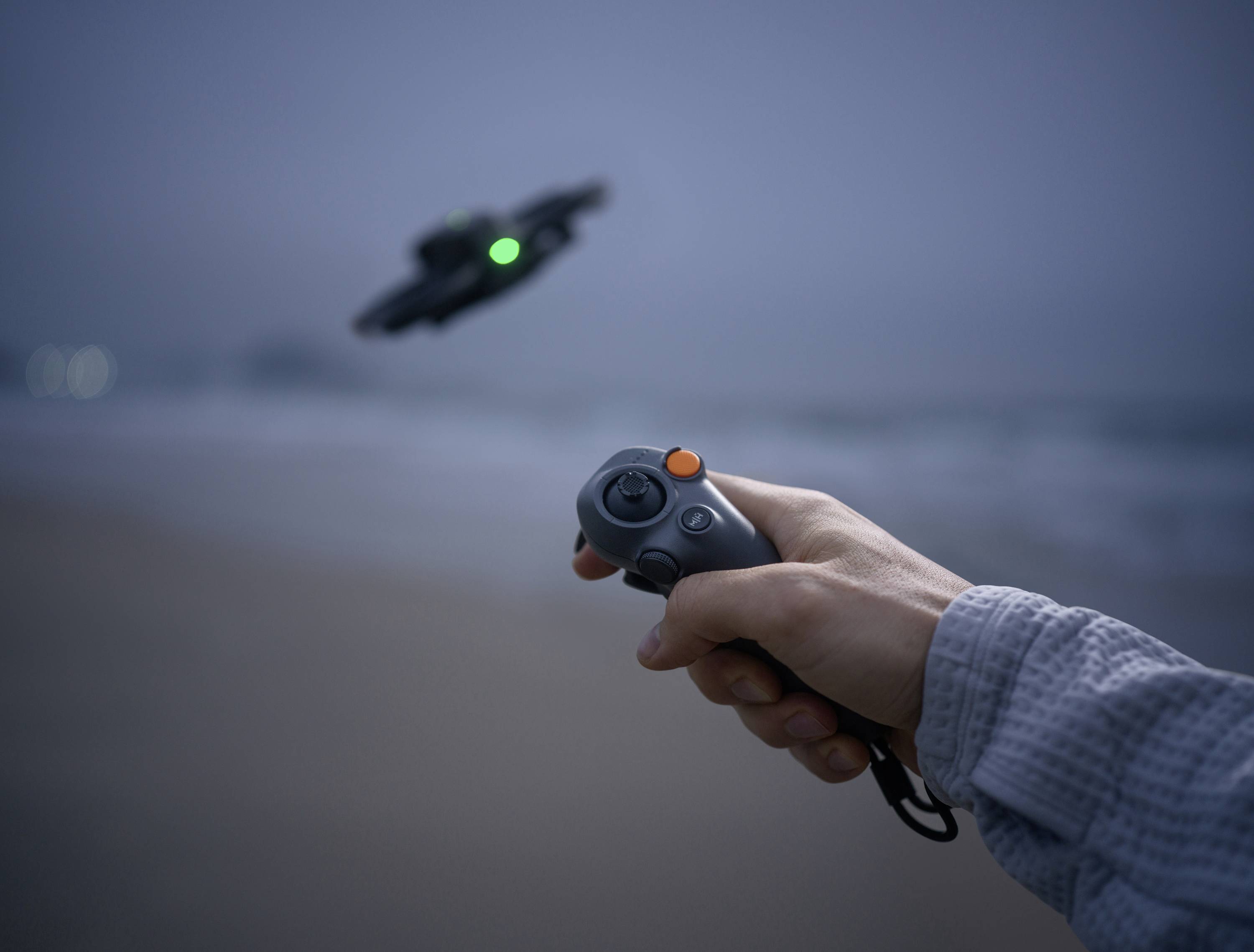 Une main pilote un drone avec une télécommande sur une plage au crépuscule. Le drone a une lumière verte et survole la mer.