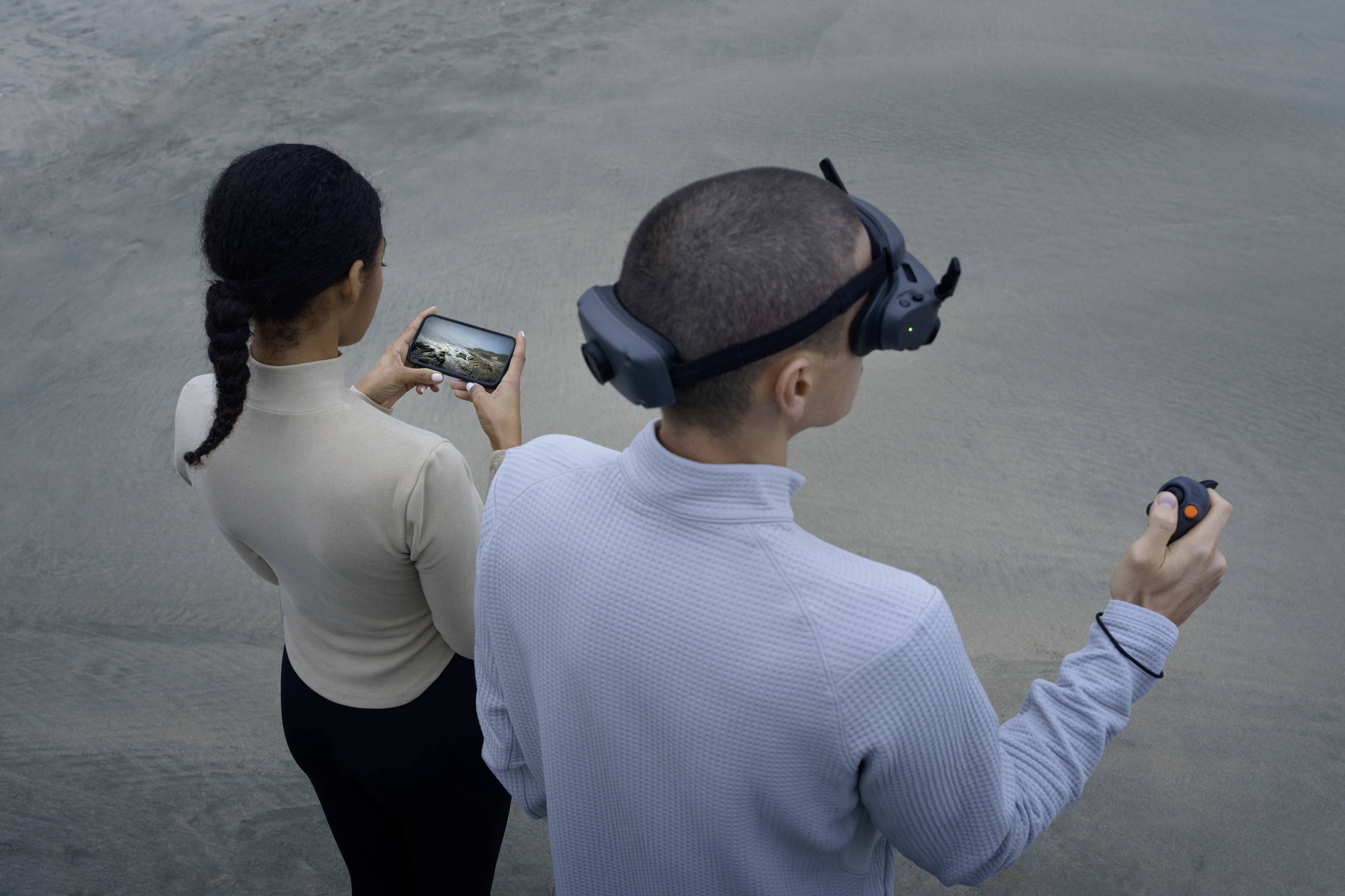 Deux personnes se tiennent sur un sol sablonneux. Une femme tient un smartphone affichant un paysage. Un homme porte un casque de réalité virtuelle et tient une manette.
