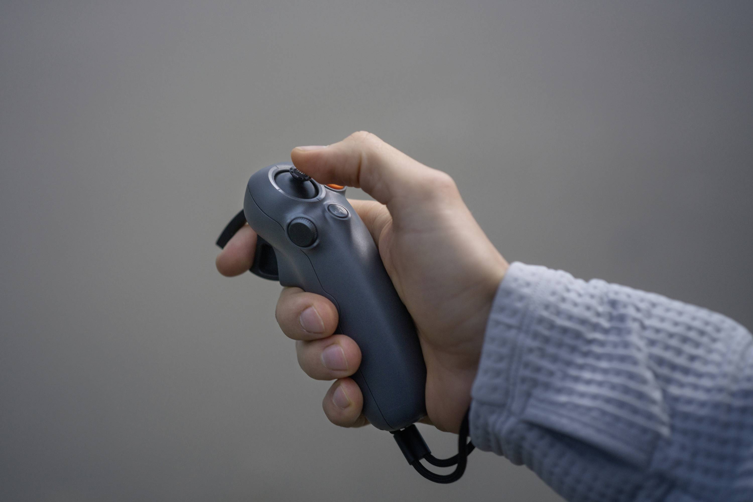 Une main tient une manette grise avec plusieurs boutons et un joystick. L'arrière-plan est flou et de couleur neutre.