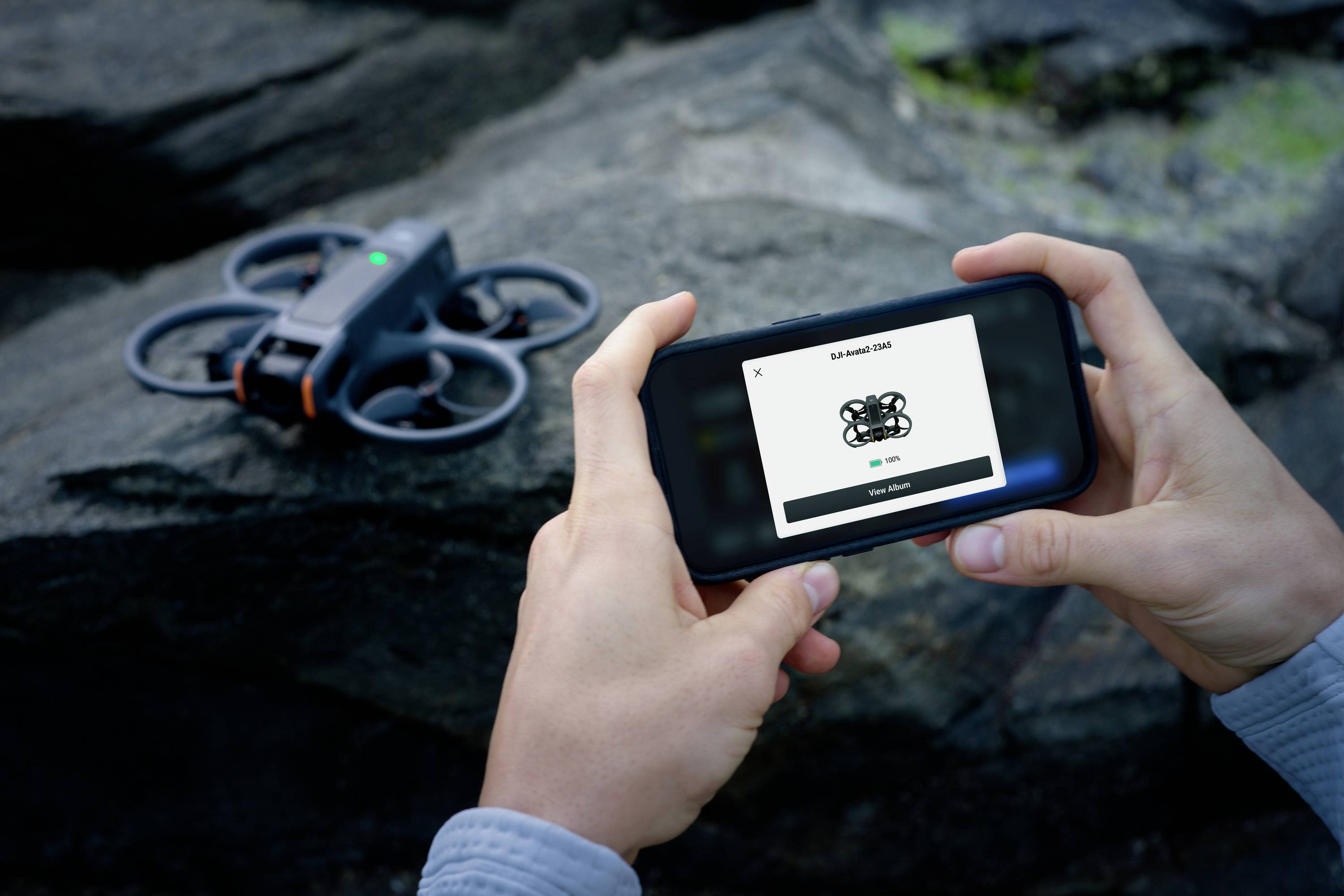 Une personne tient un smartphone avec une interface de contrôle de drone. Un petit drone est posé sur un rocher en arrière-plan, prêt à décoller.