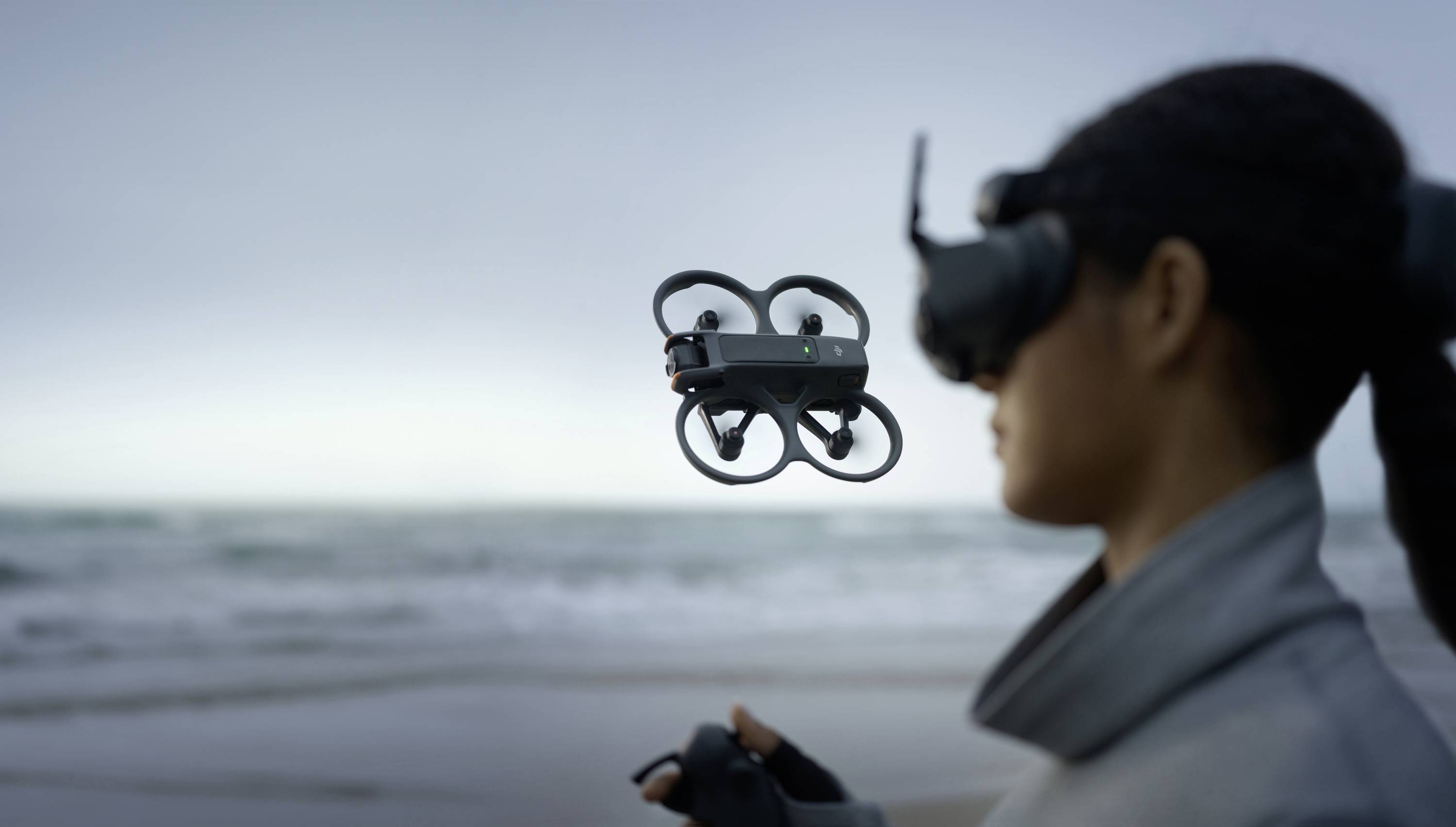 Une personne se tient sur la plage et pilote un petit drone en vol stationnaire devant elle, à l'aide d'une télécommande et d'un casque de réalité virtuelle.
