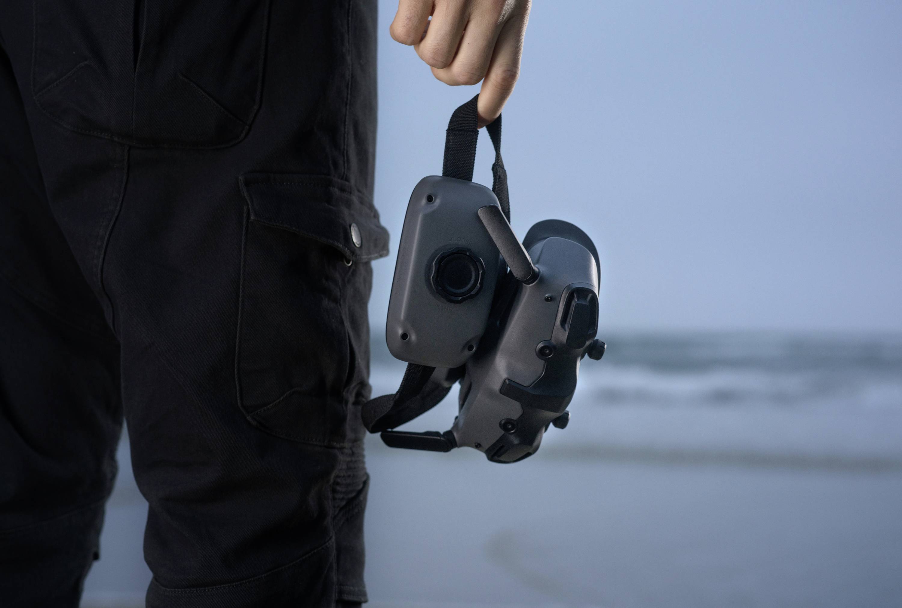 Une personne tient un casque de réalité virtuelle à la main alors qu'elle se trouve sur une plage. En arrière-plan, on aperçoit une mer floue.