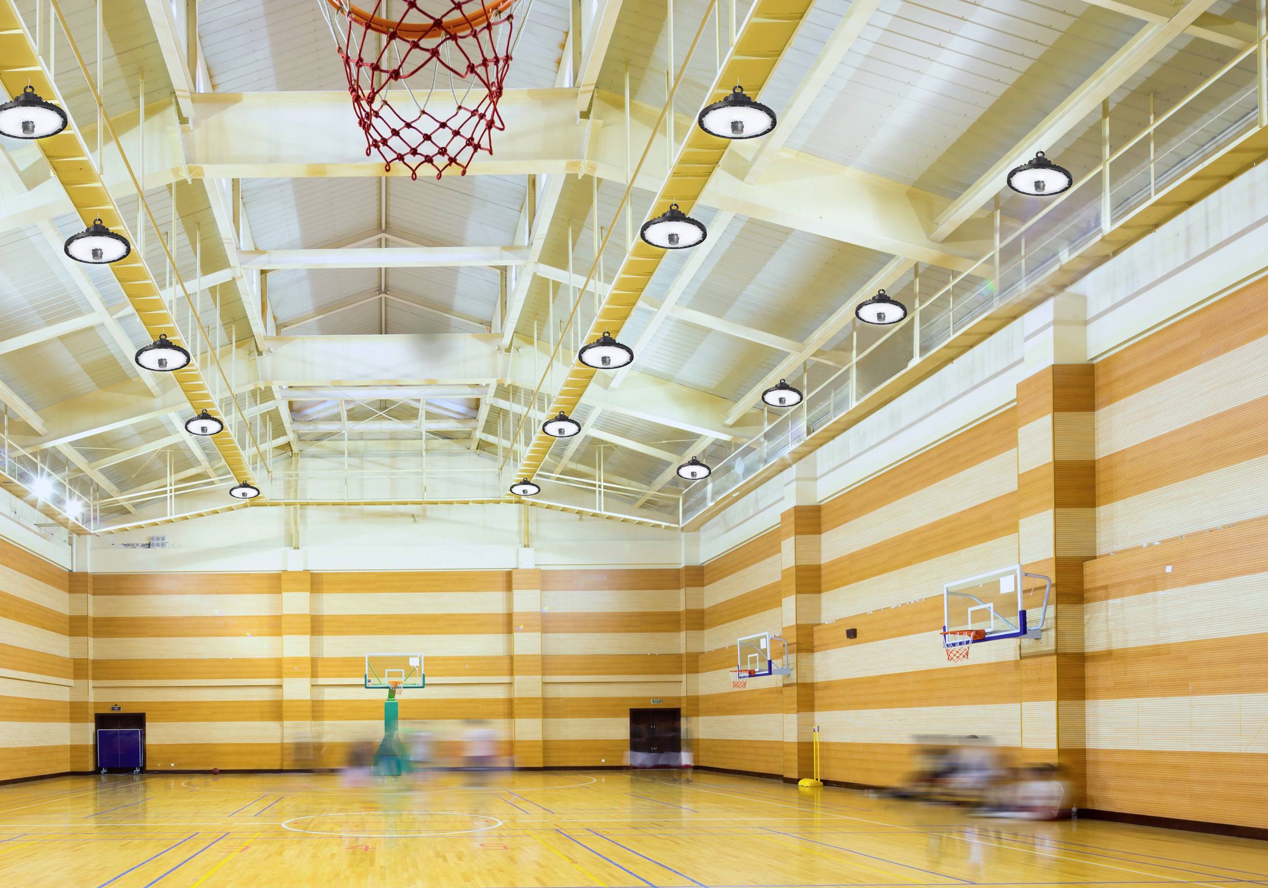 Une salle de sport vide avec un sol en bois et des paniers de basket sur les côtés. Le plafond est haut, avec des poutres apparentes et des lumières.