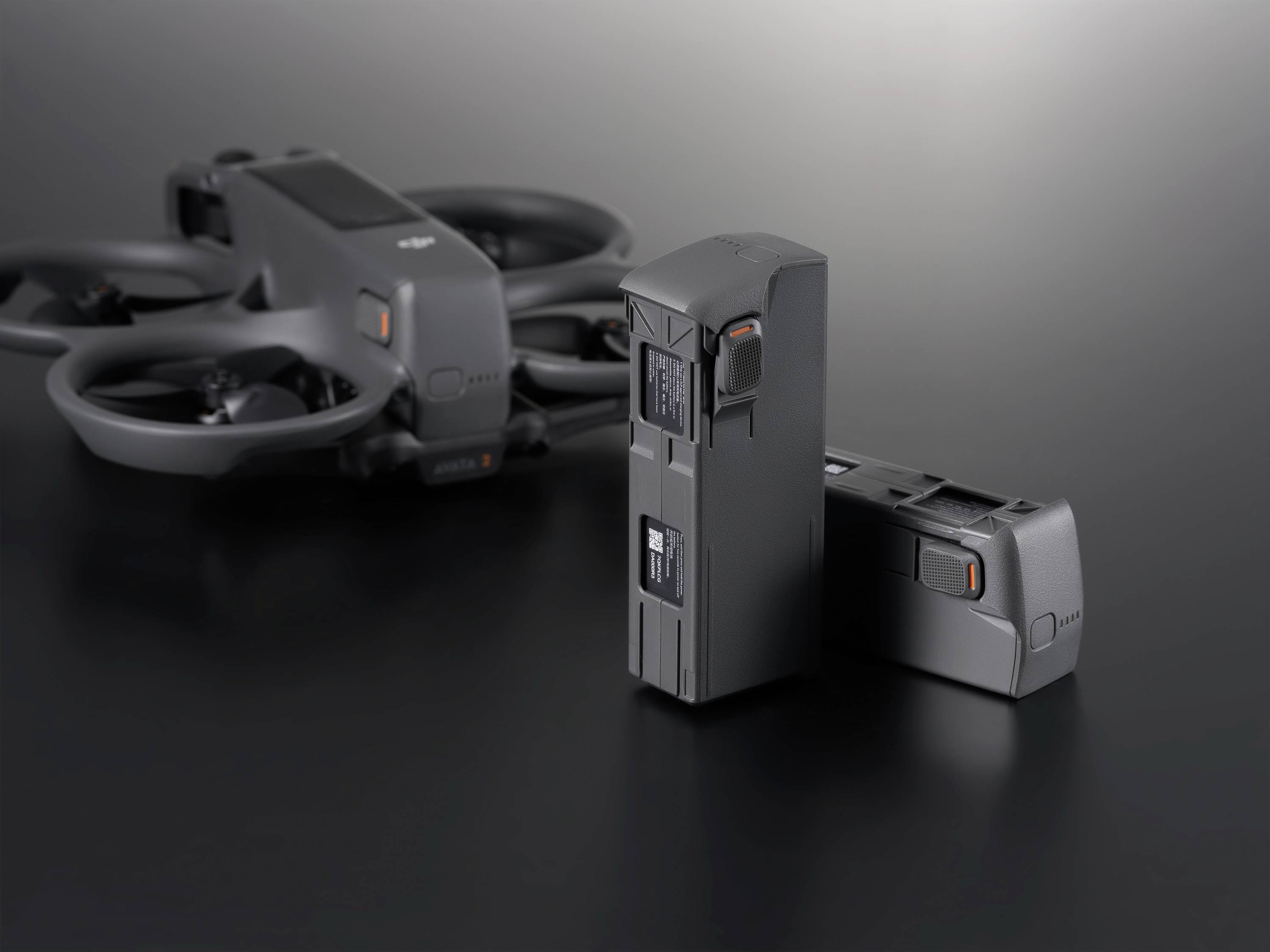 Un drone noir et deux batteries sont posés sur une table sombre. Le drone est équipé de protections d'hélices.