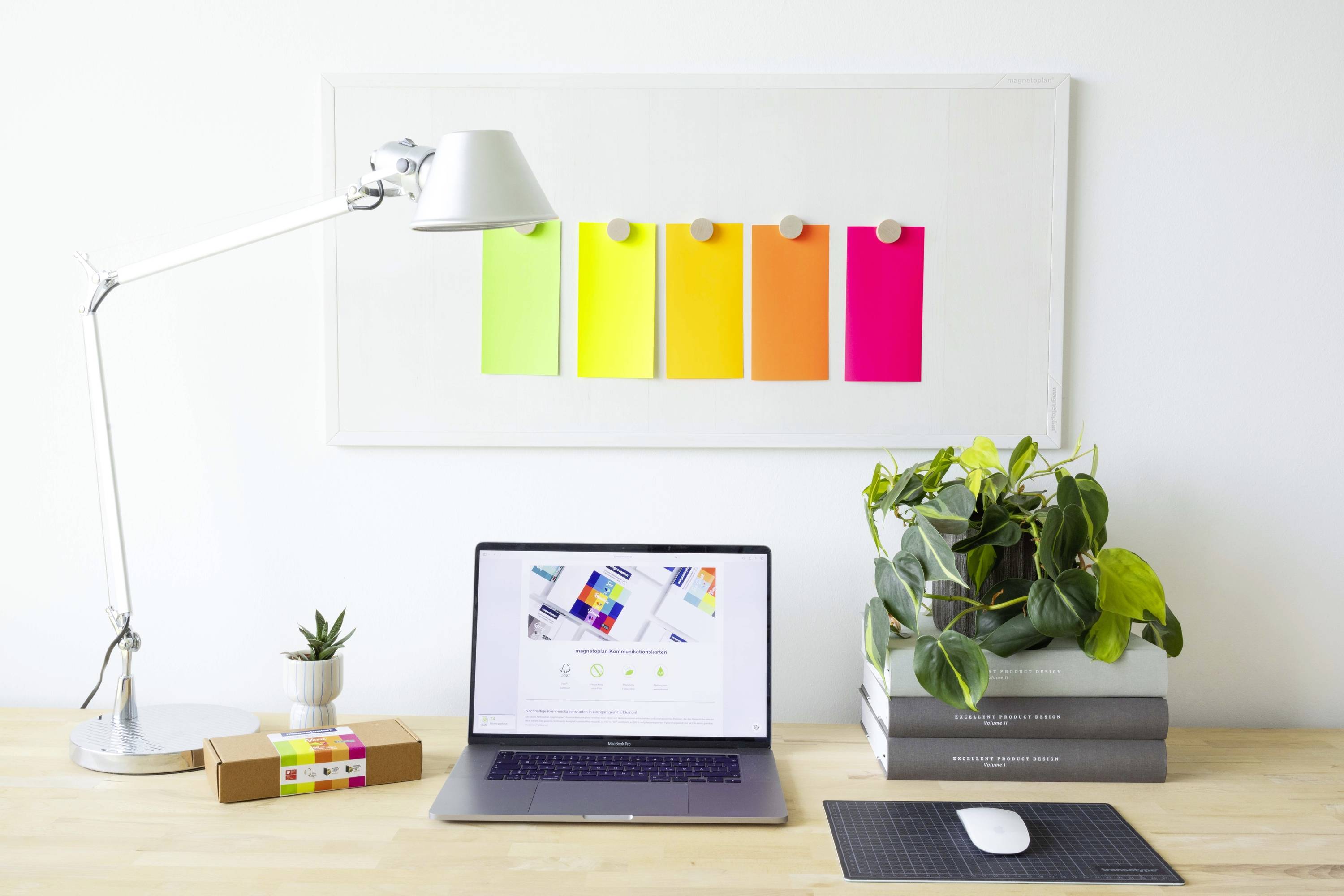 Un bureau blanc avec un ordinateur portable ouvert, une plante verte, un petit cactus, une lampe et des cartes colorées sur le tableau d'affichage.