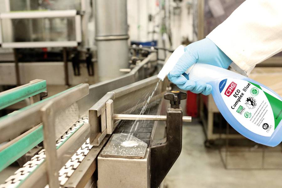 Une personne portant des gants bleus vaporise un produit de nettoyage sur un tapis roulant dans un environnement industriel.