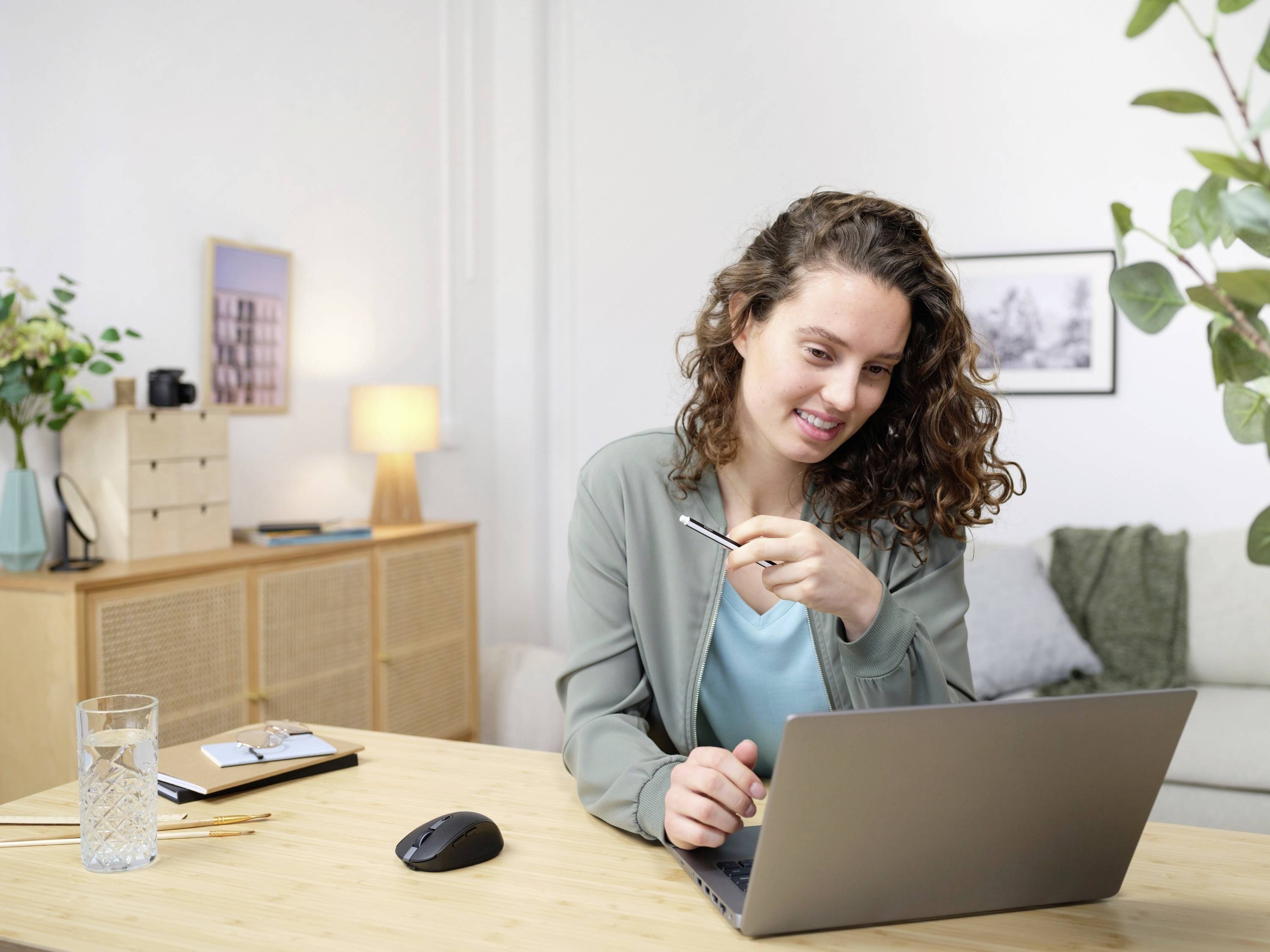 Femme aux cheveux bouclés assise à une table, utilisant un ordinateur portable, souriant. Des plantes et des tableaux sont visibles en arrière-plan.