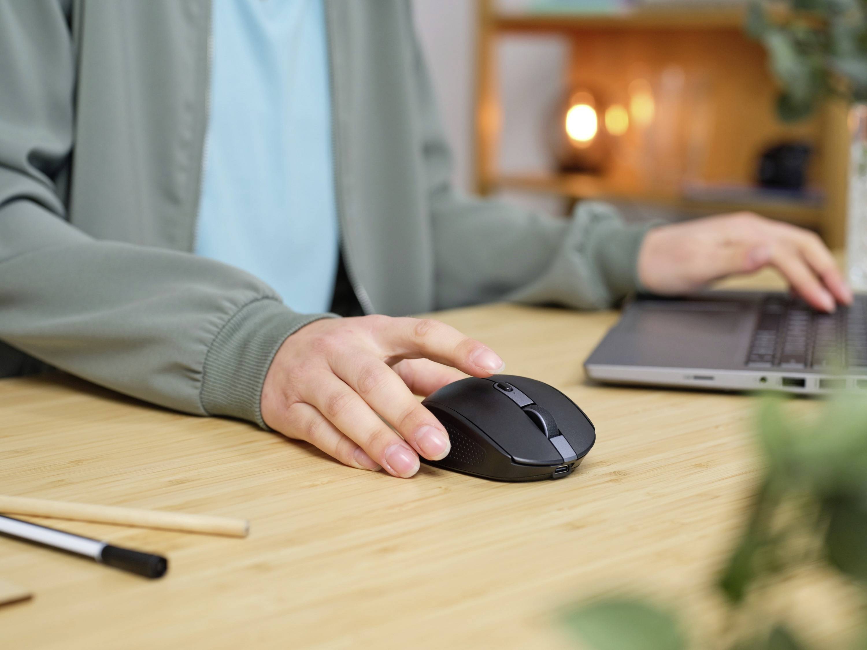 Une personne travaille sur un ordinateur portable, utilise une souris sans fil, est assise à une table avec des stylos au premier plan ; l'arrière-plan montre des étagères.