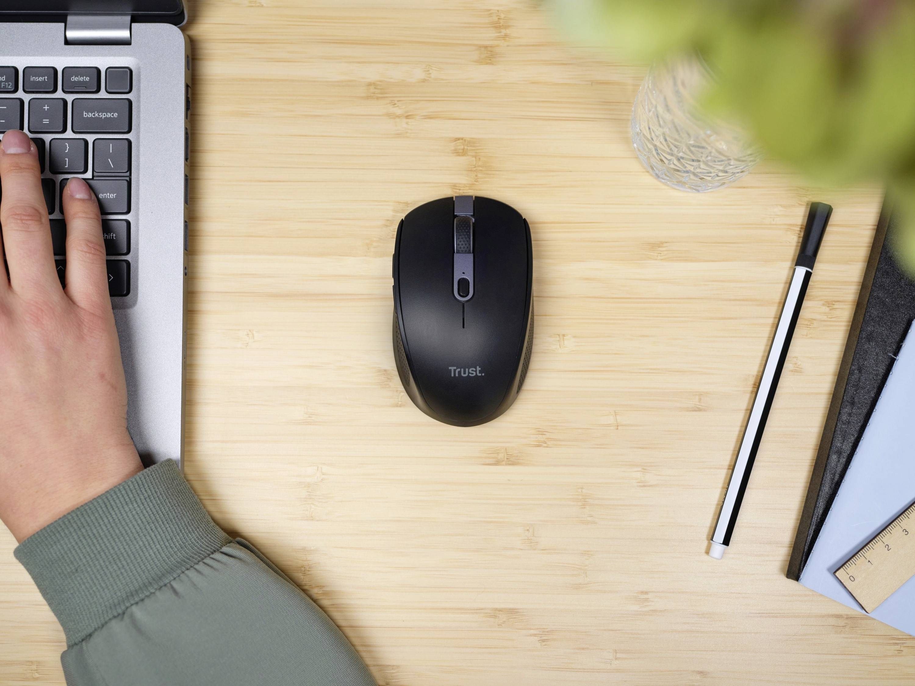 Un ordinateur portable avec une main sur le clavier, à côté une souris sans fil noire sur une table en bois, avec un stylo et un carnet de notes à proximité.
