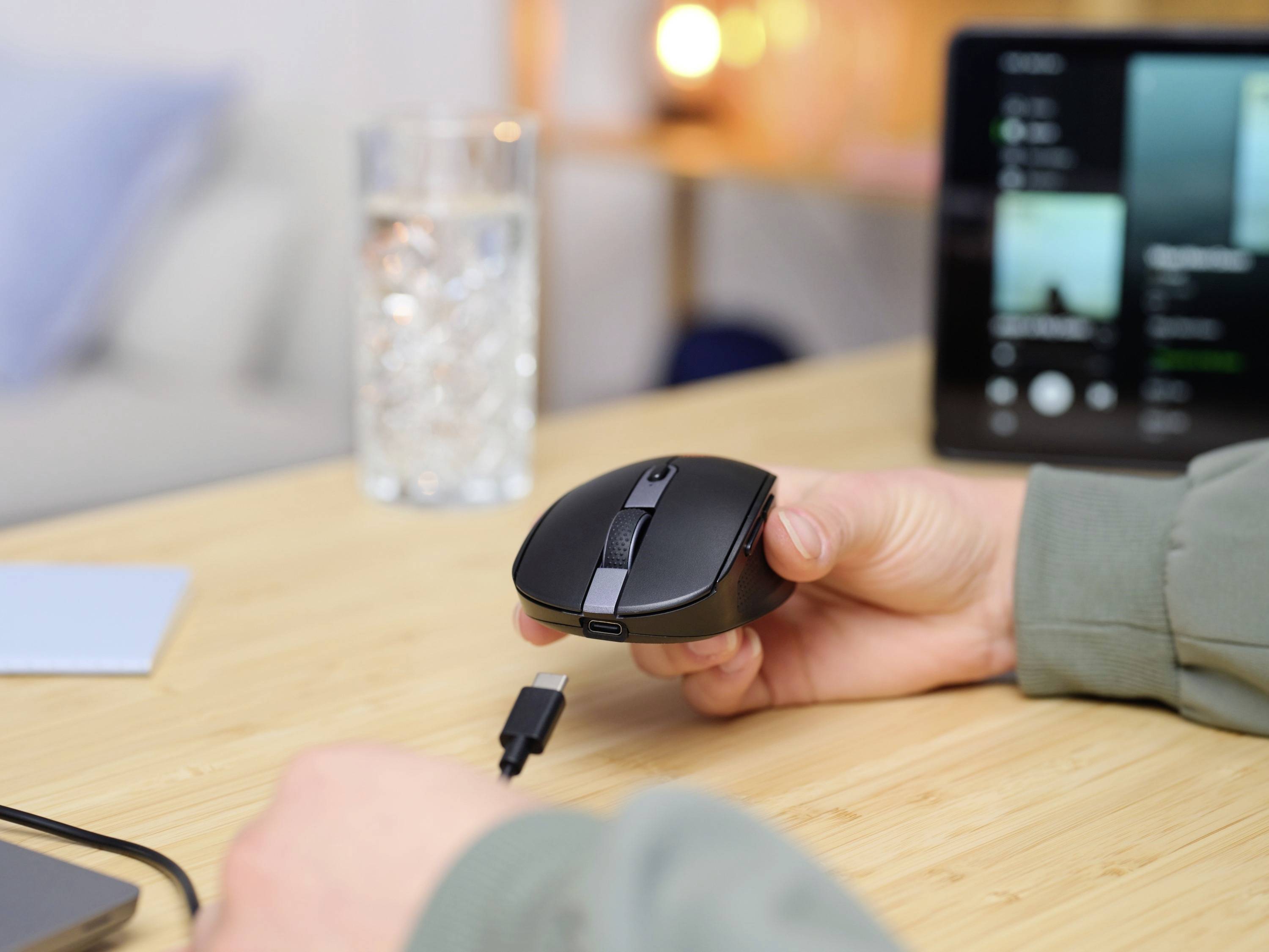 Une personne tient une souris d'ordinateur noire, prête à être branchée avec un câble USB. Un verre d'eau et un écran sont visibles en arrière-plan.