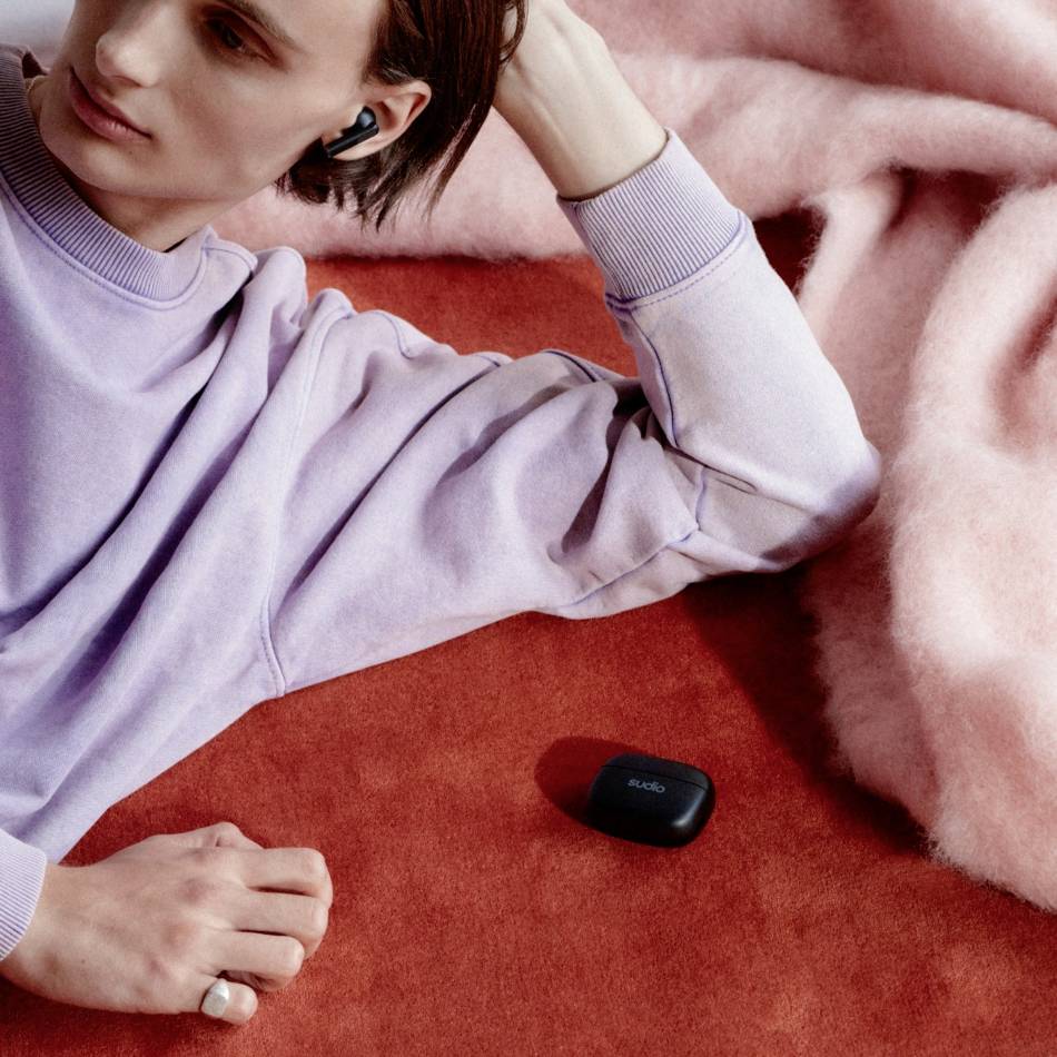 Une personne est allongée de manière détendue sur un tapis rouge, portant un pull violet et des écouteurs, avec une coque noire à côté, sur un fond rose.