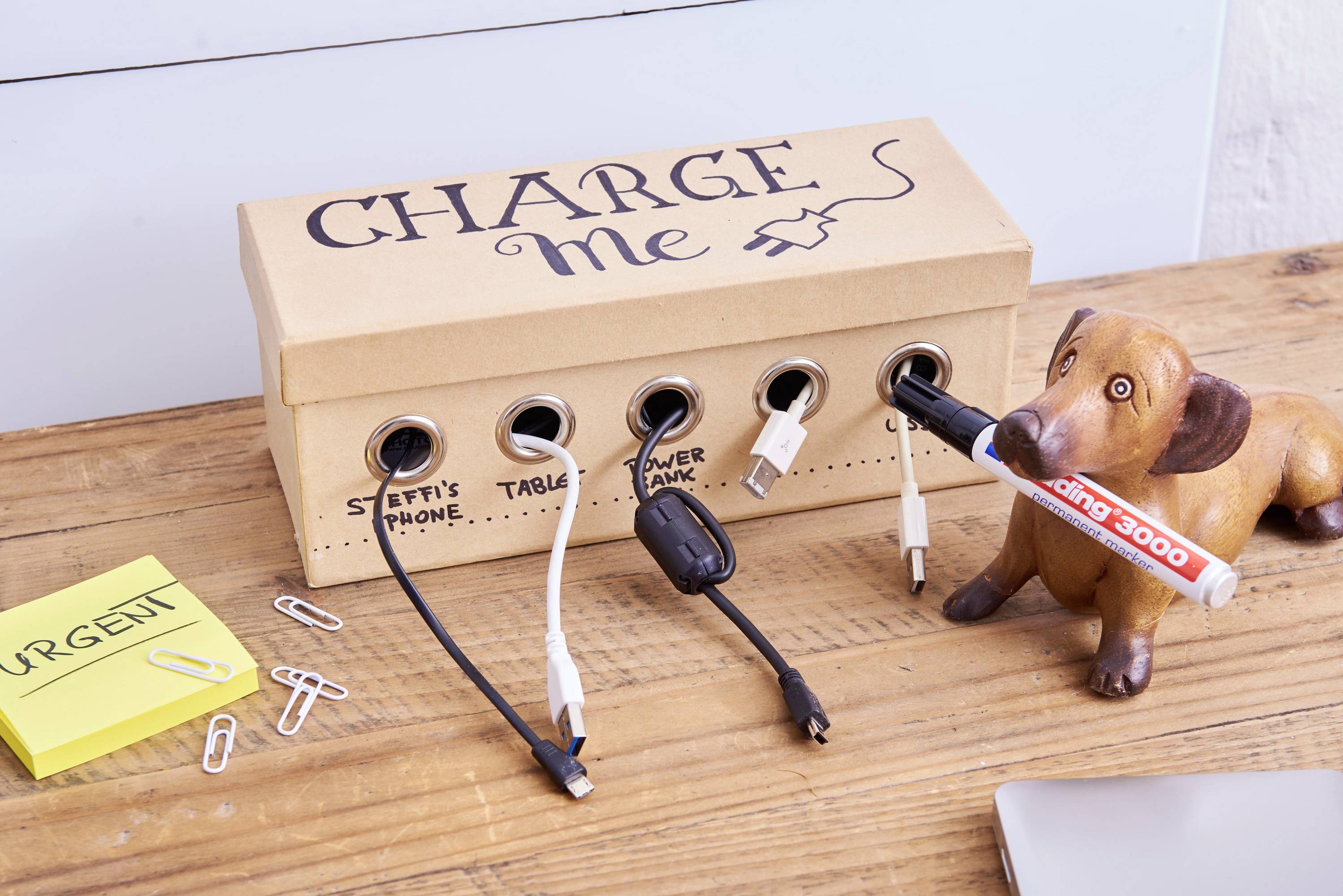 Une boîte en carton étiquetée 'Charge Me', avec quatre ports de chargement pour 'Téléphone de Steffi', 'Tablette', 'Batterie externe'. À côté se trouve un jouet pour chien avec un stylo dans la gueule.