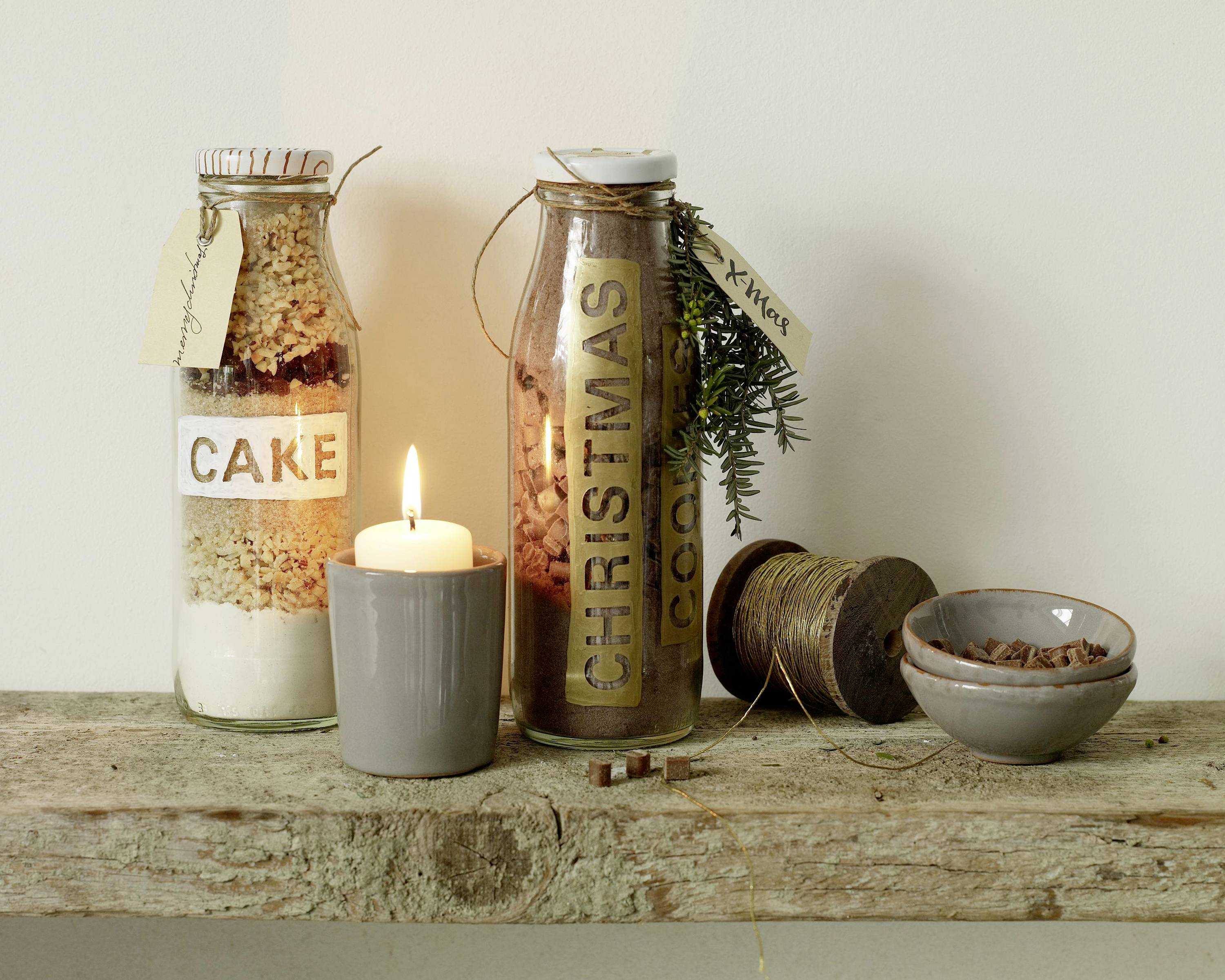 Deux bocaux en verre contenant des ingrédients pour gâteau et biscuits, étiquetés 'Cake' et 'Christmas Cookies', sont posés sur une étagère en bois à côté d'une bougie allumée.