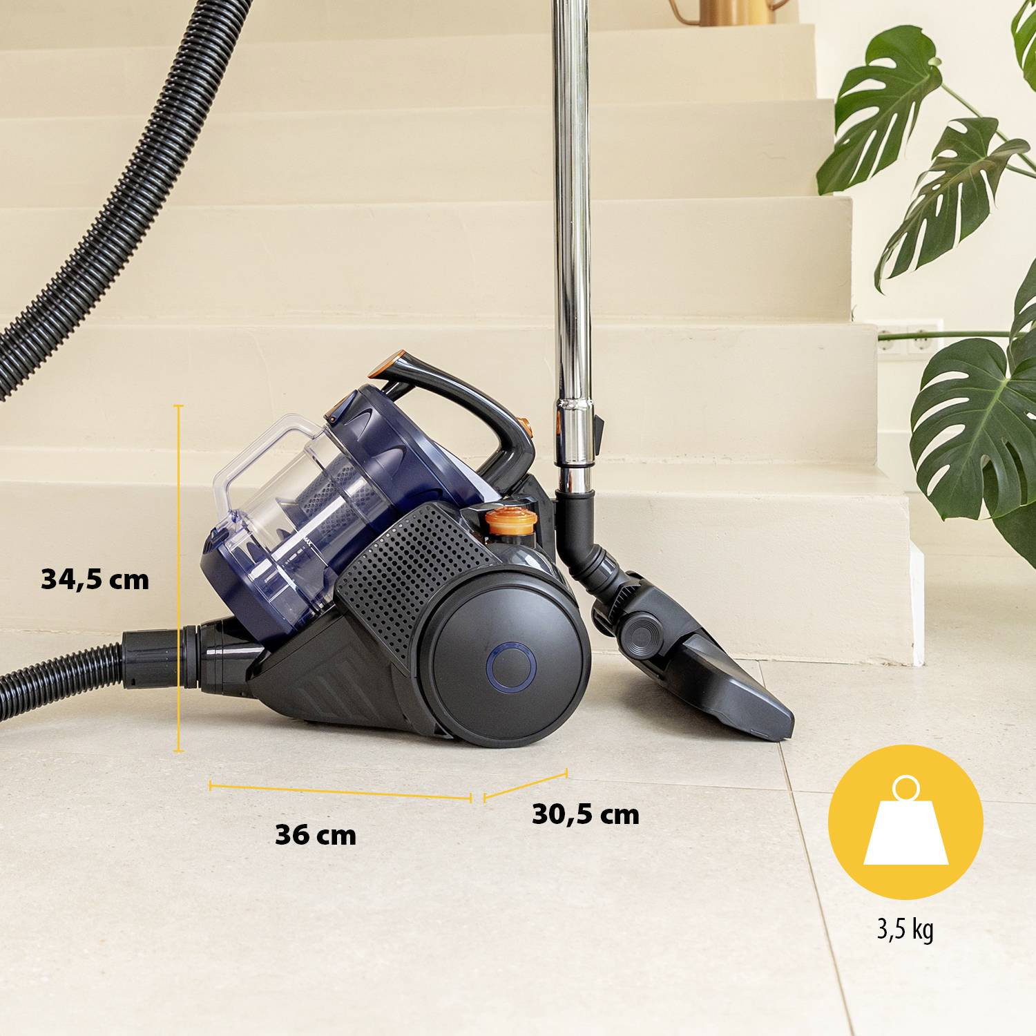 Aspirateur avec tuyau et accessoires, posé sur un carrelage devant un escalier. Dimensions : 34,5 cm de hauteur, 36 cm de profondeur, 30,5 cm de largeur. Poids 3,5 kg.